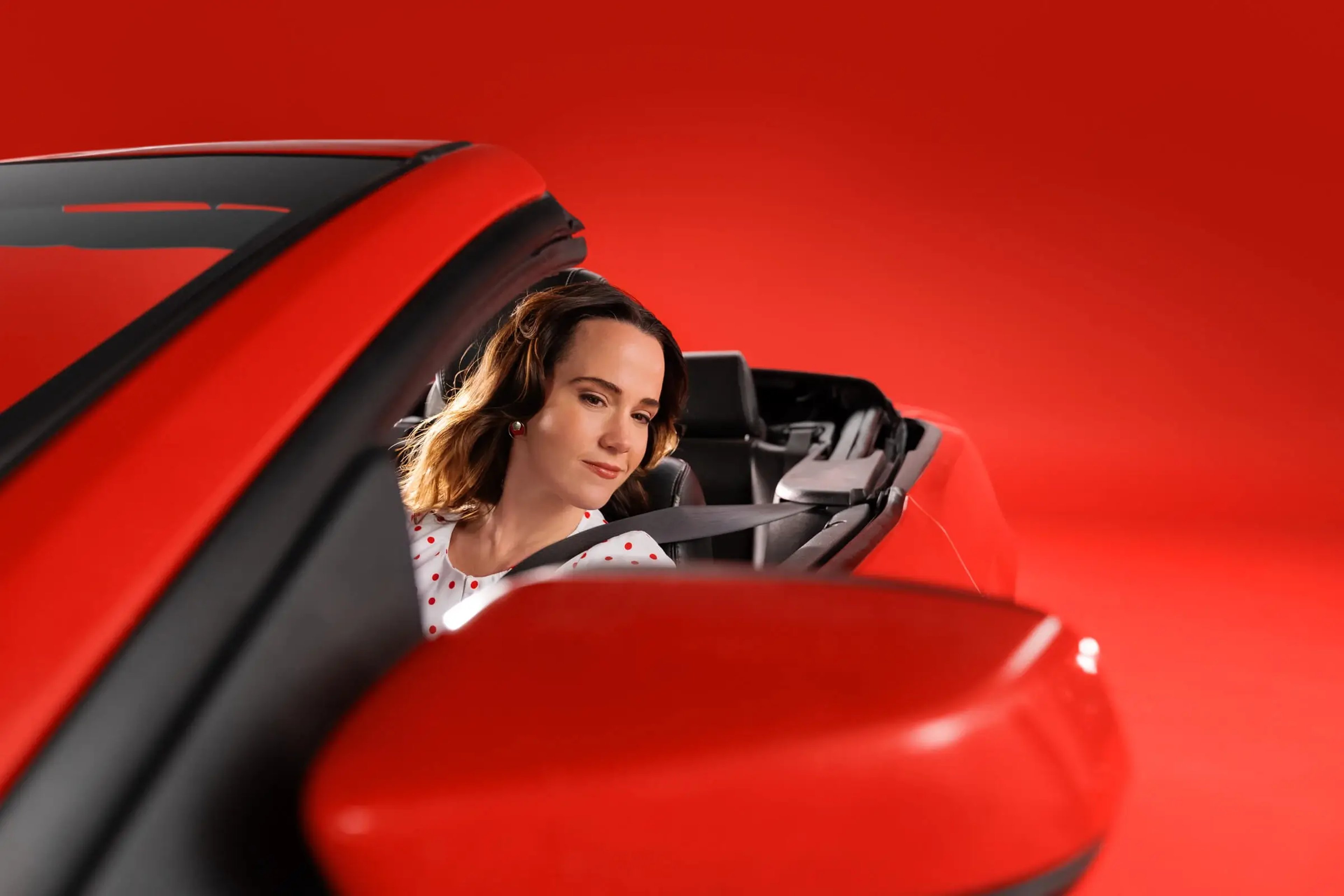 A woman in a white dress with red polka dots sitting in the driver's seat of a red convertible car against a solid red studio backdrop.