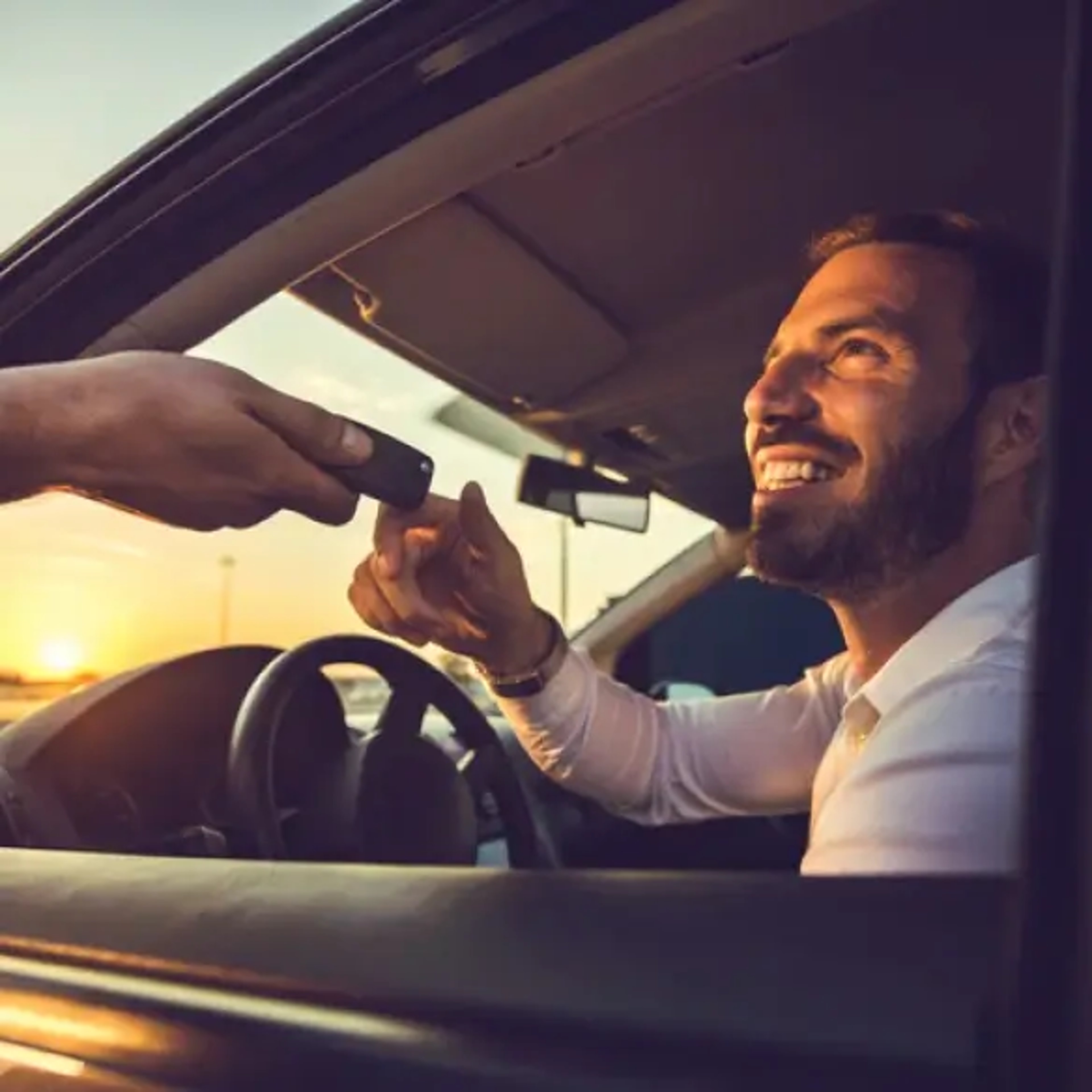 A smiling man sits in the driver’s seat of a car during sunset, reaching out to accept a car key from another person.