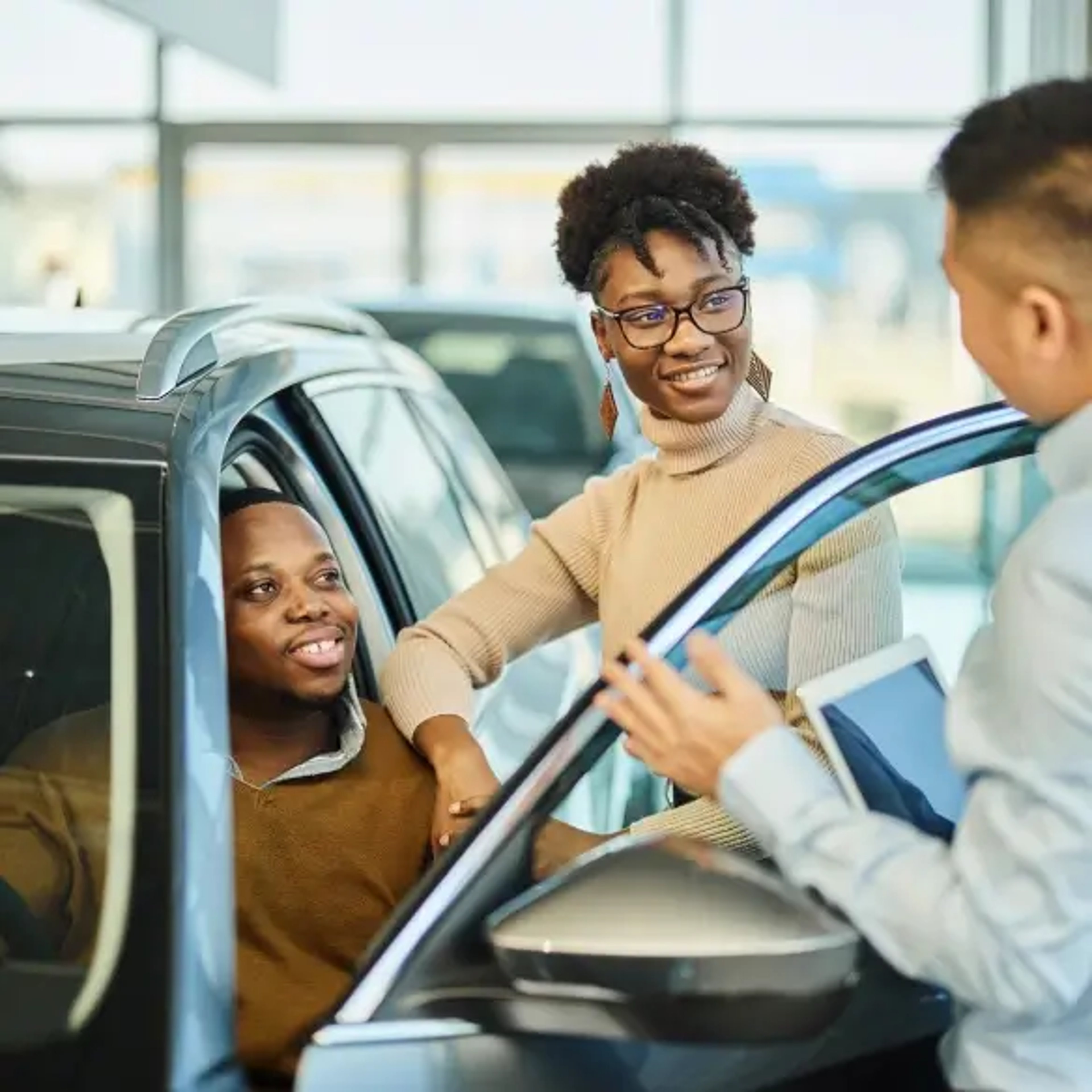 A smiling couple interacts with a professional standing by their car in a dealership setting. The man is seated inside the car while the woman leans on the open door, engaging in a discussion. The professional, holding a tablet, appears to be explaining or presenting information about the vehicle.