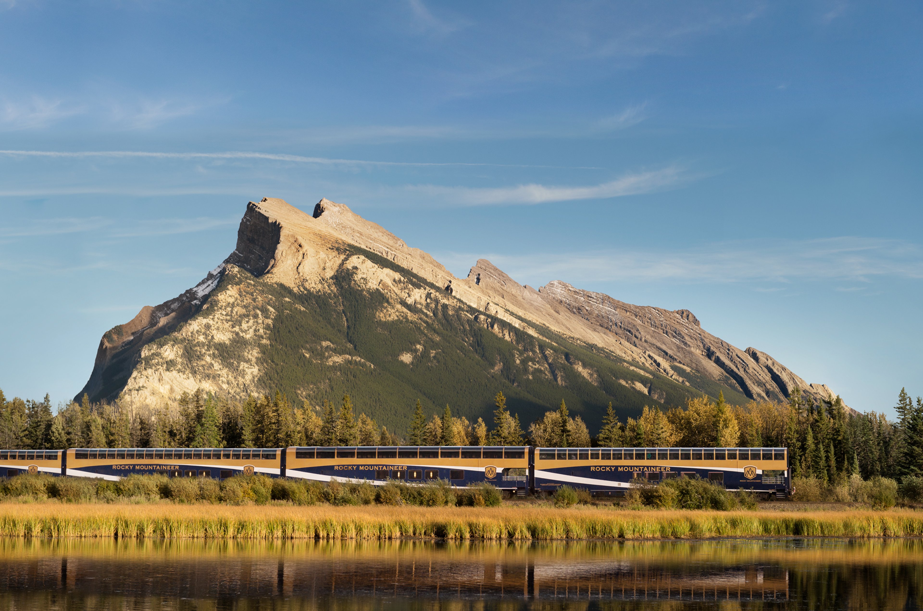 Rocky Mountaineer train passing along a river with a massive mountain in the background 