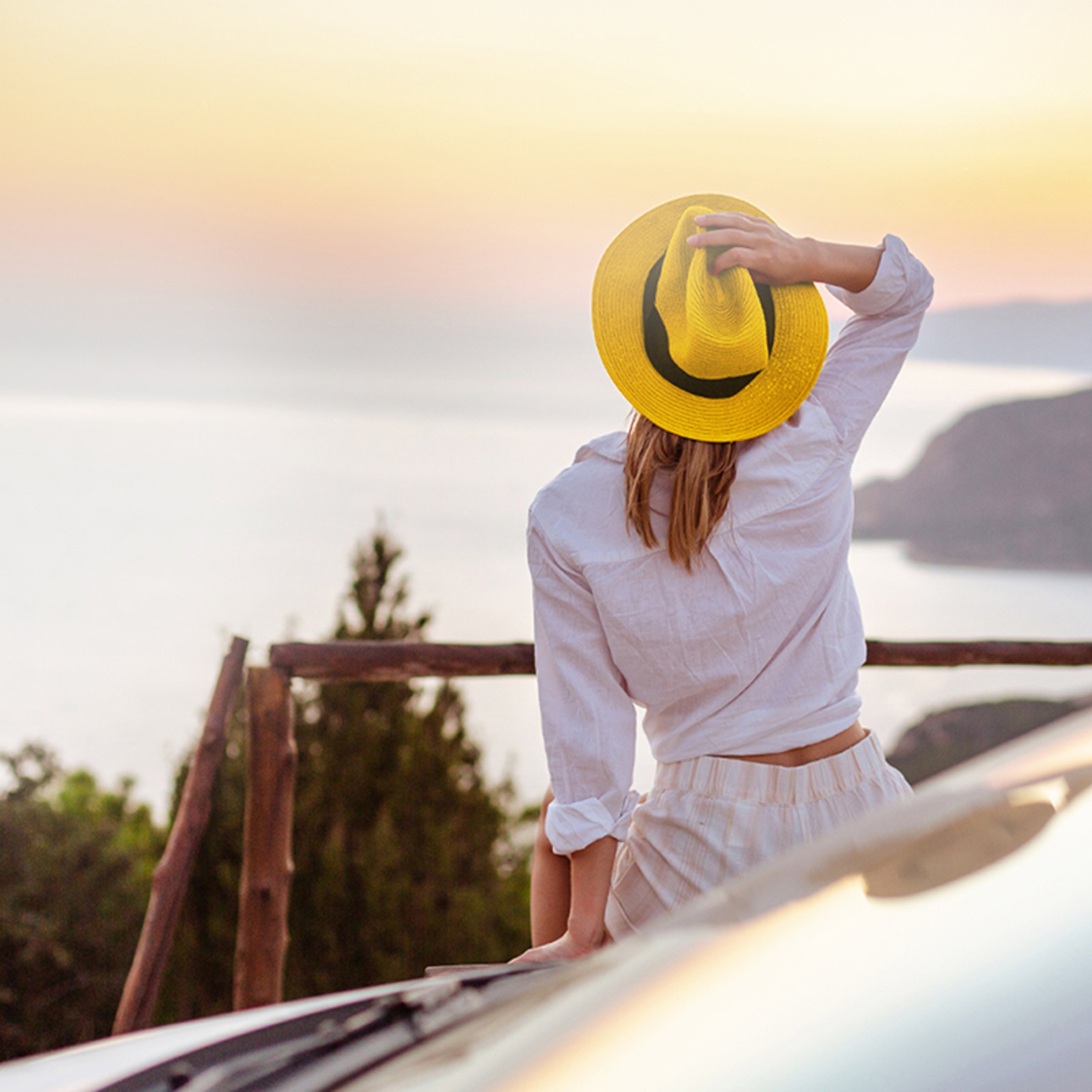 Photo from behind of woman standing by car looking out over a scenic coastal view while holding a Hertz yellow fedora hat on her head