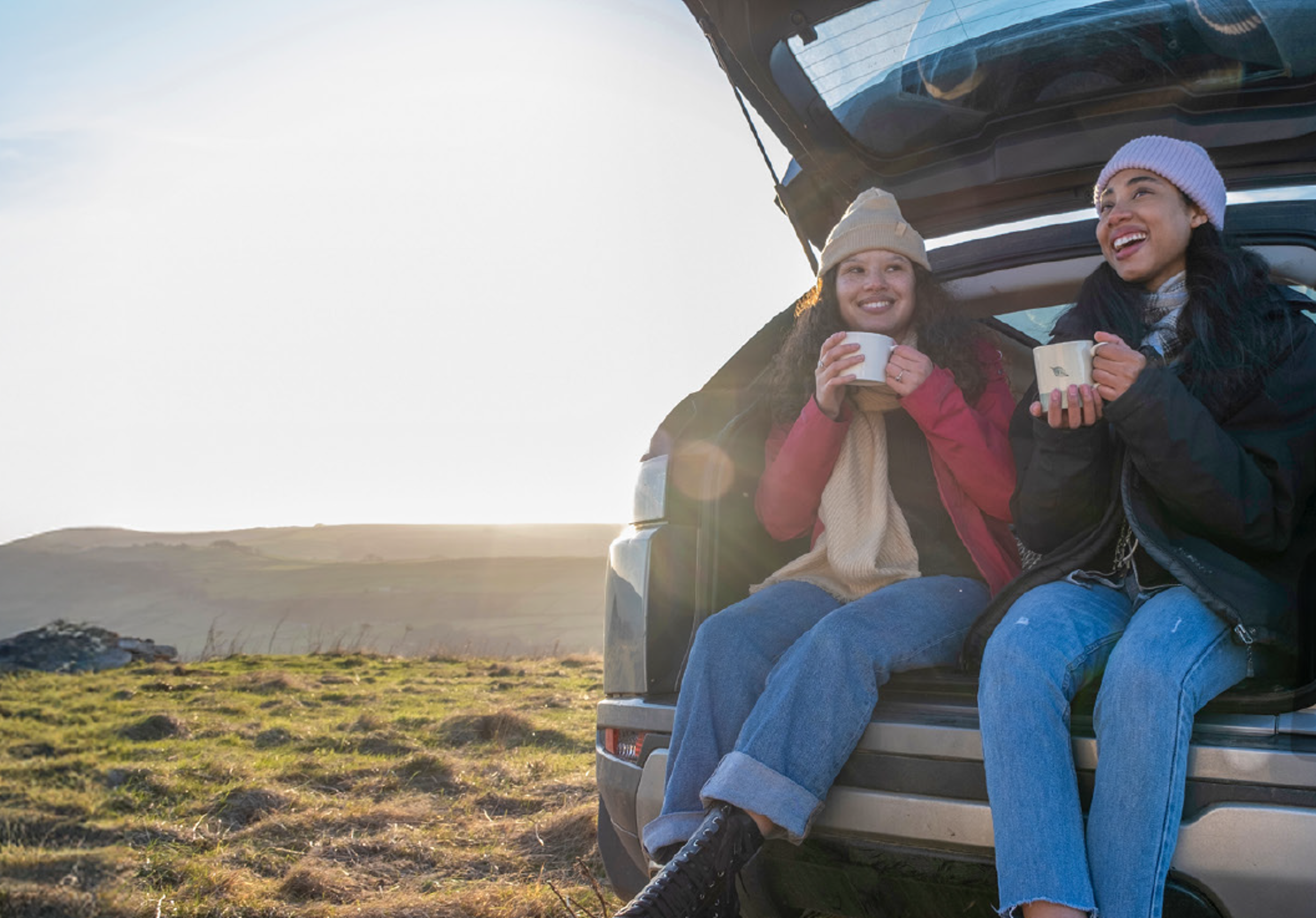 Two young women enjoying cup of coffee in trunk of rental SUV