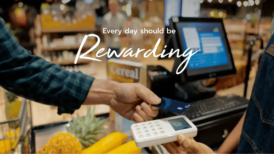 A shopper uses a credit card at a grocery store checkout, with fresh produce visible in the cart. The text overlay reads, "Every day should be Rewarding," emphasizing the benefits of earning rewards on everyday purchases.