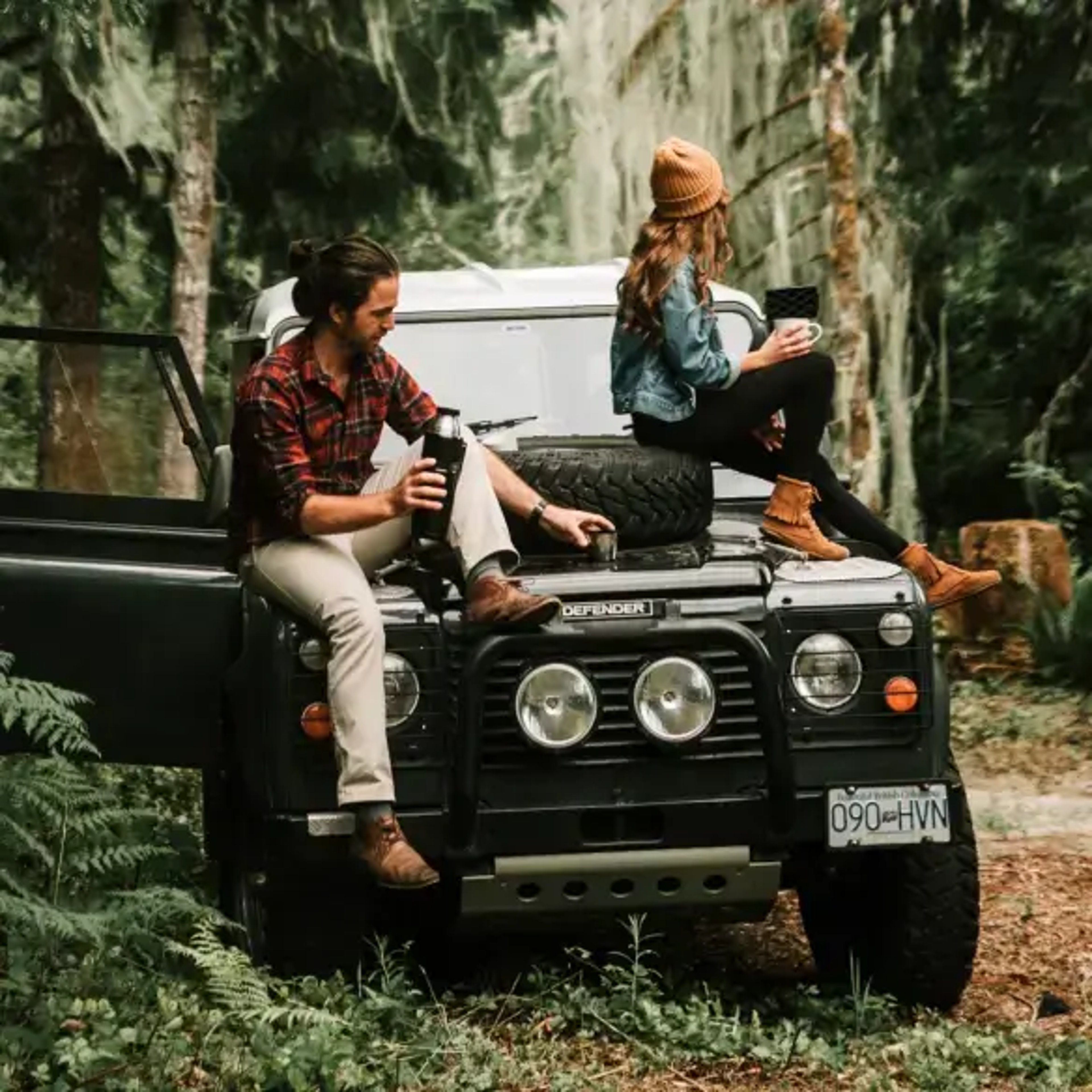 A man and woman enjoy a moment in a forested area, sitting on a rugged SUV with an open door. The man, dressed in a plaid shirt, holds a thermos, while the woman, wearing a knit hat, sits on the hood holding a mug.