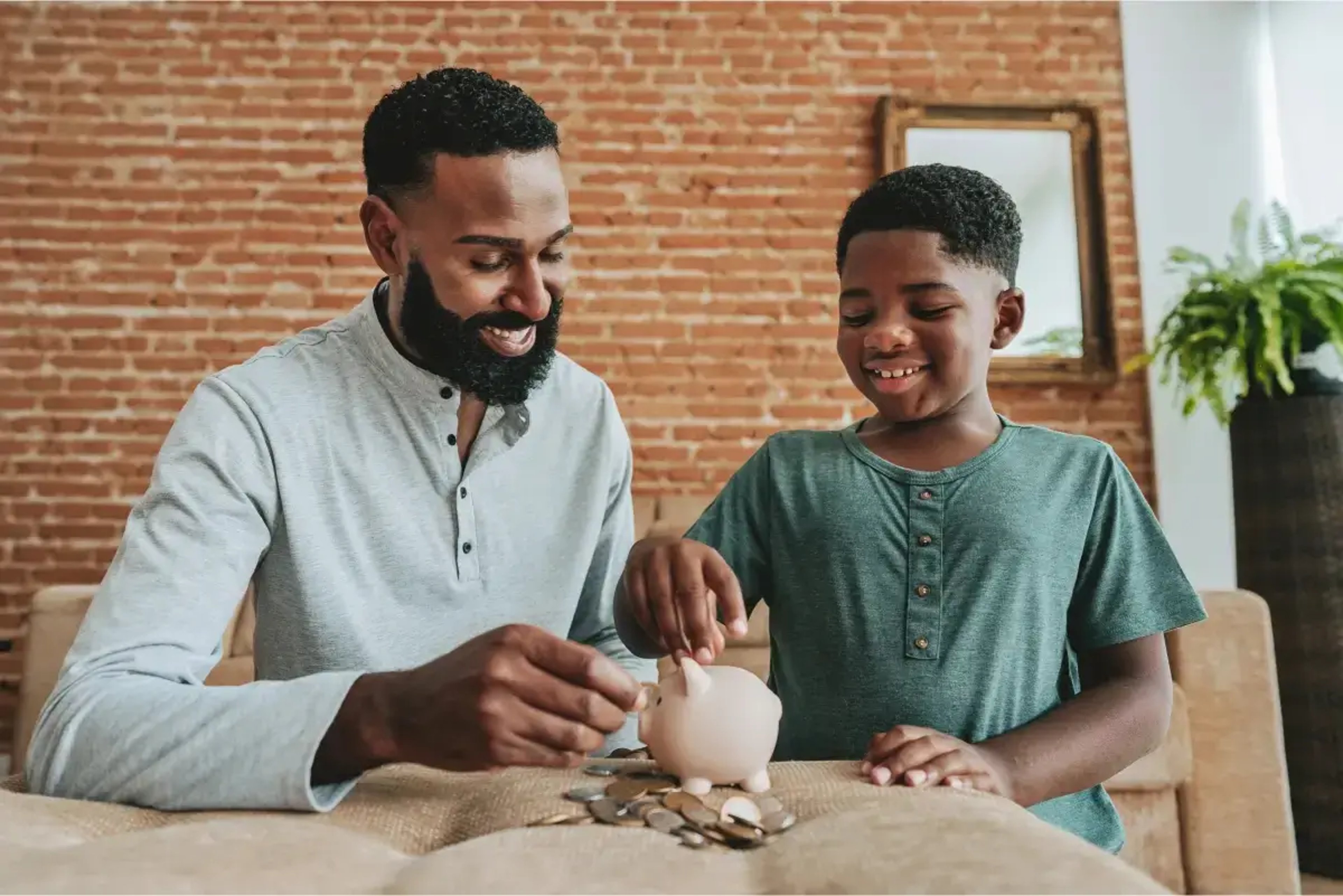 A father and son happily engage in saving money together at home, placing coins into a pink piggy bank on a table. The background features a cozy living room with a brick wall, a framed mirror, and greenery.