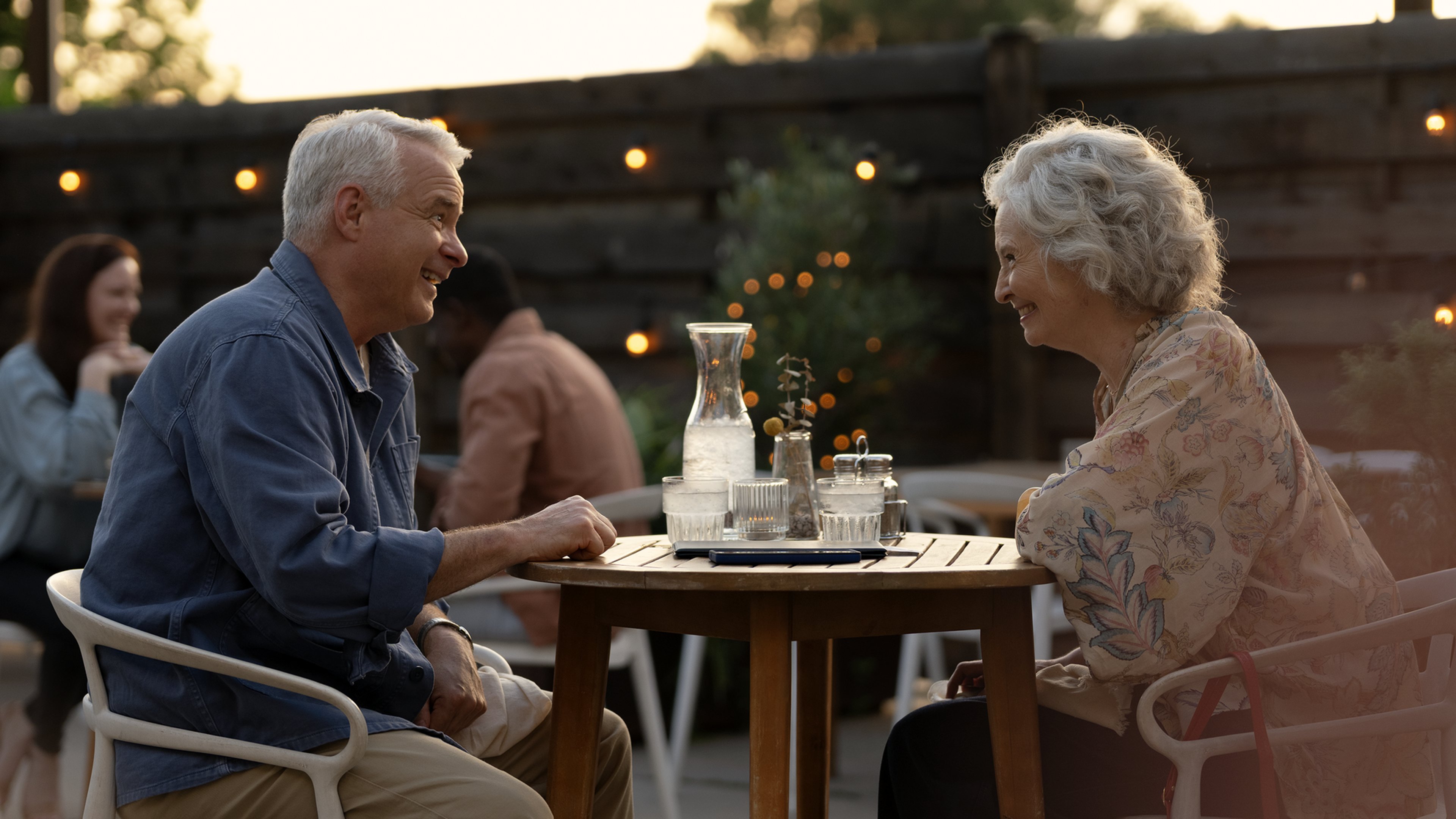 An outdoor cafe setting with two individuals seated at a table facing each other, surrounded by wooden furniture and soft lighting.