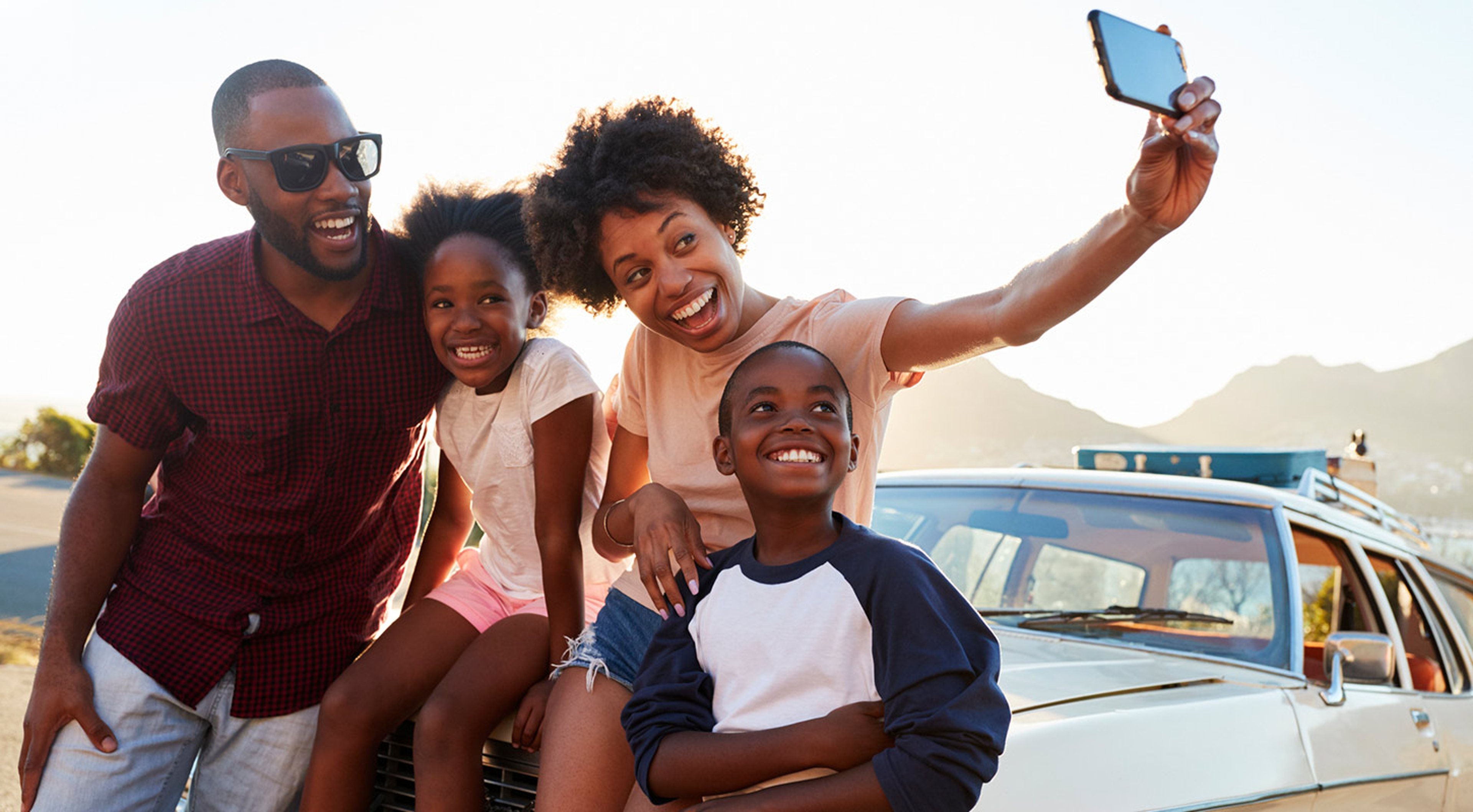 A family of four taking a selfie next to a car with mountains in the background.