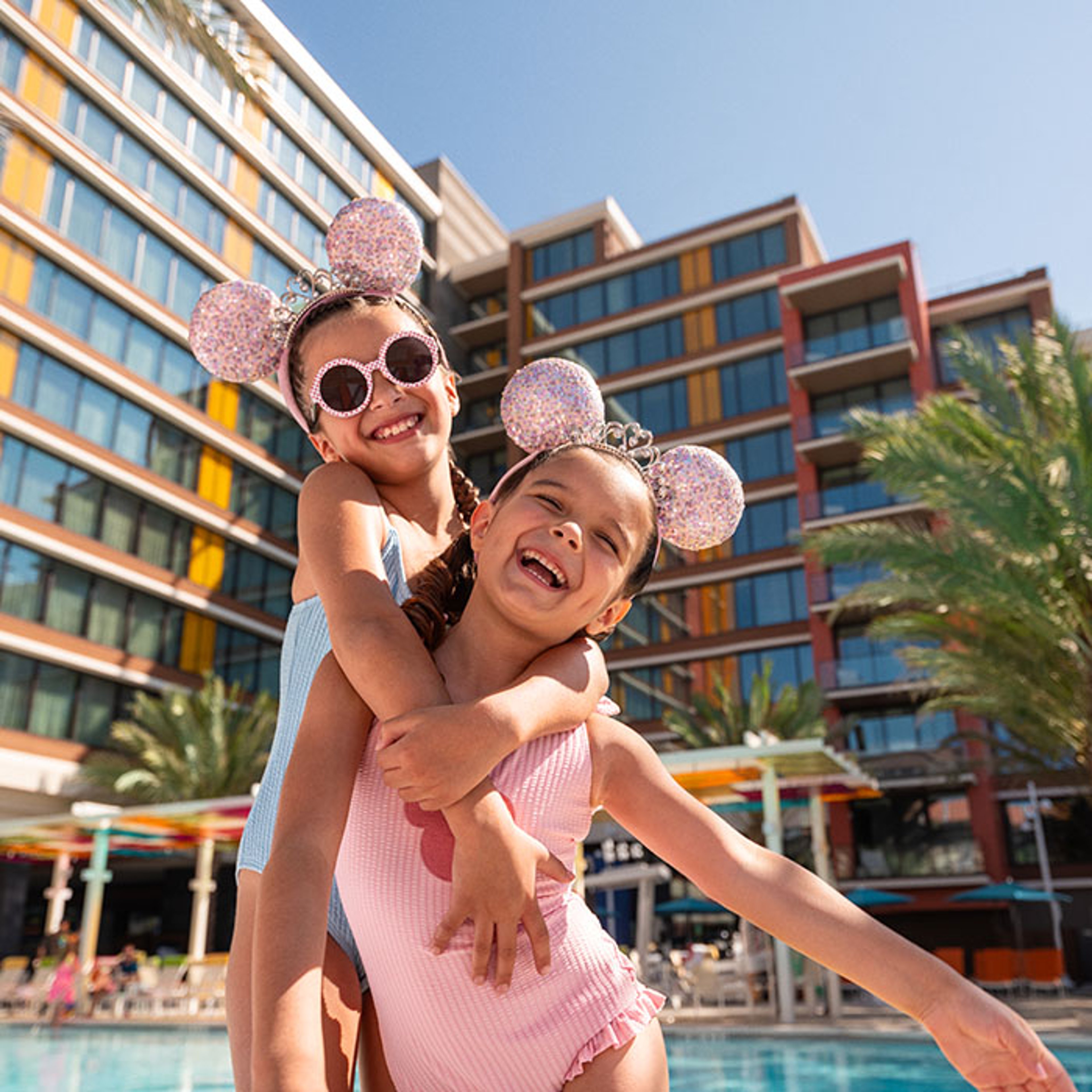 Two smiling children wearing sparkly mouse-ear headbands hug and laugh beside a resort pool with colorful hotel buildings in the background.