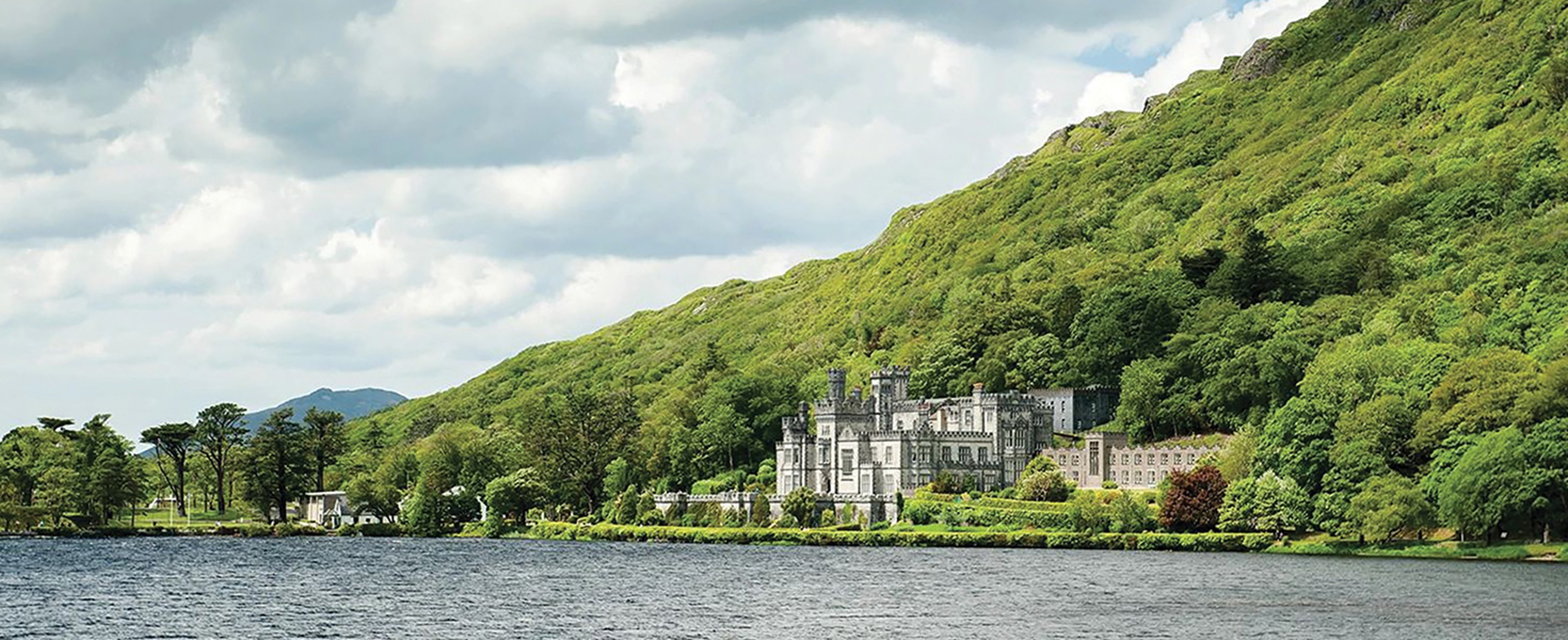 The image shows Kylemore Abbey, a historic neo-Gothic castle and Benedictine monastery nestled against a wooded mountainside beside Pollacappul Lough in Connemara, County Galway, Ireland