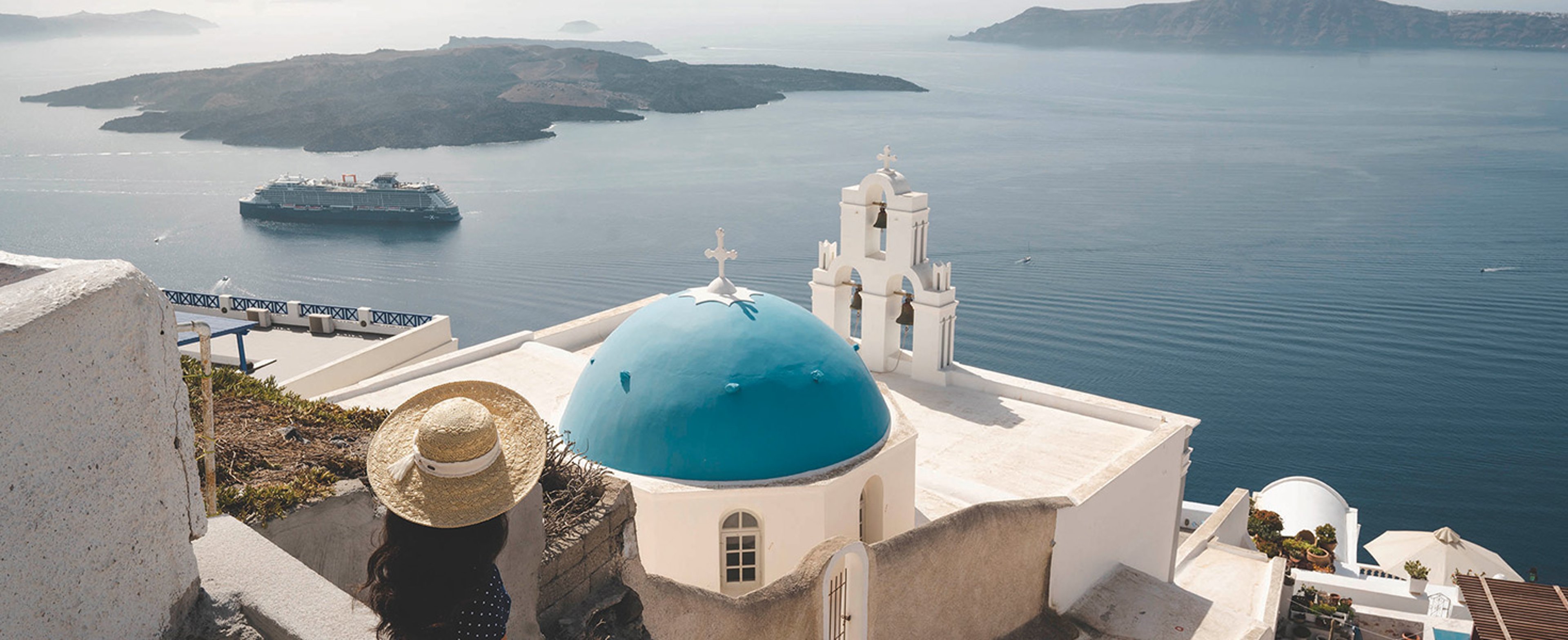 The image shows the iconic Three Bells of Fira church overlooking the caldera in Santorini, Greece with a Celebrity cruise ship sailing in the background.