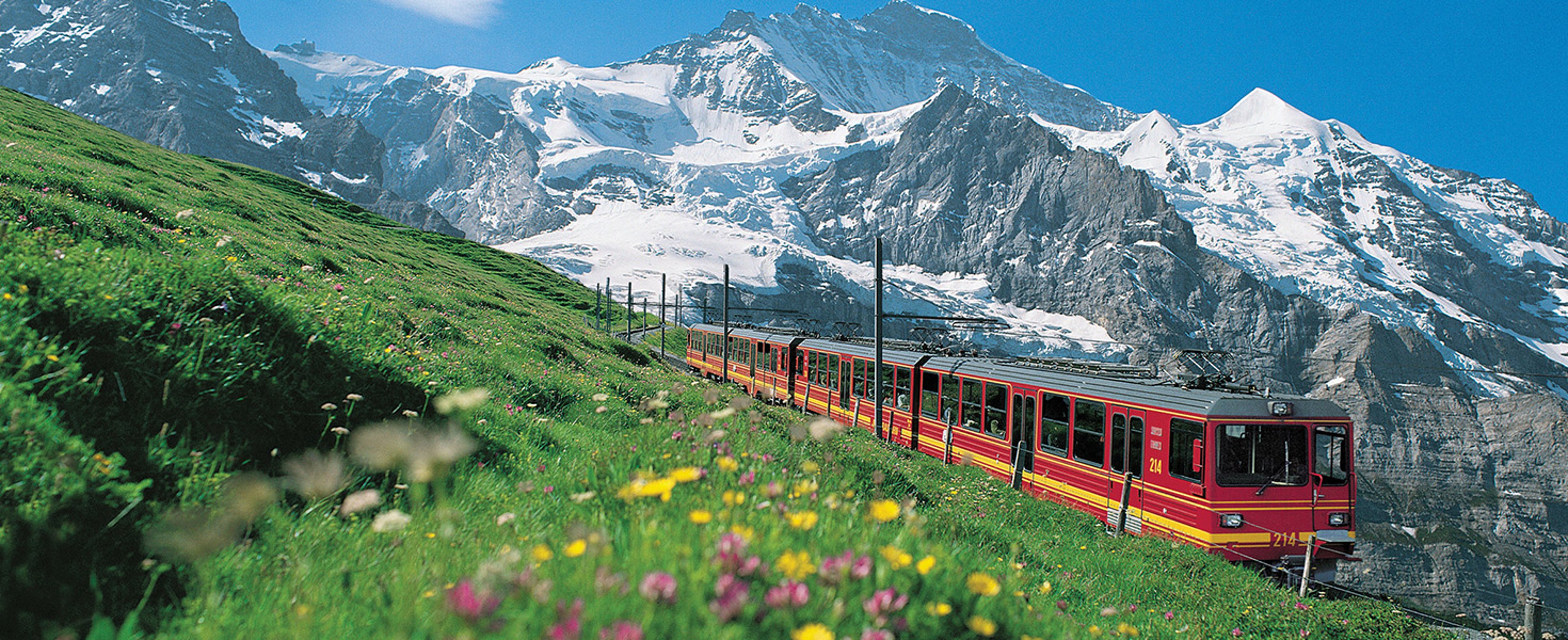 The image shows a red regional train traveling on an alpine slope with snow-capped mountains in the background, appearing to be part of the famous Jungfrau Railway system in the Swiss Alps
