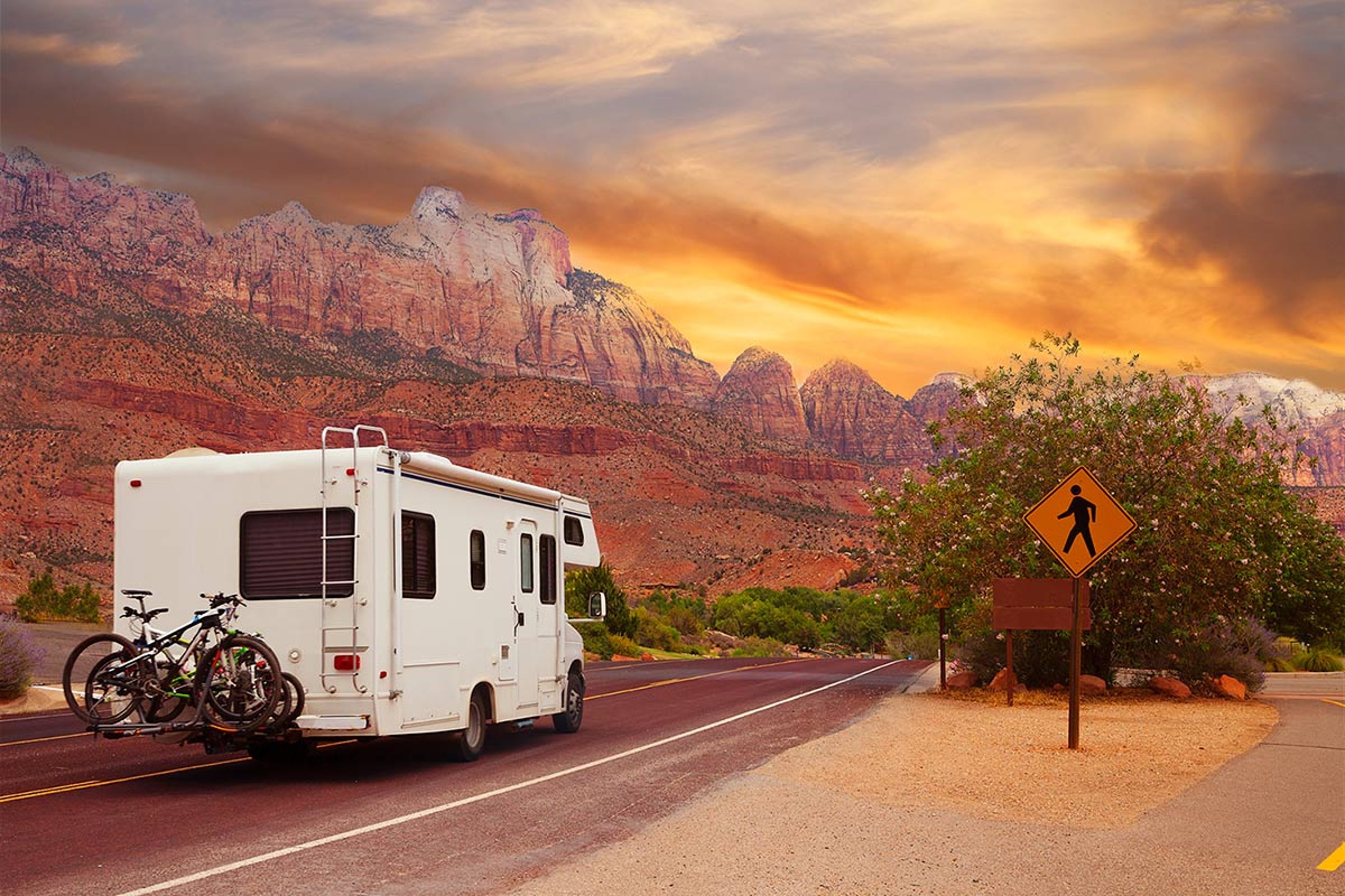 White RV with bicycles on the back driving along a road through a desert canyon at sunset, with dramatic rock formations and a pedestrian sign.