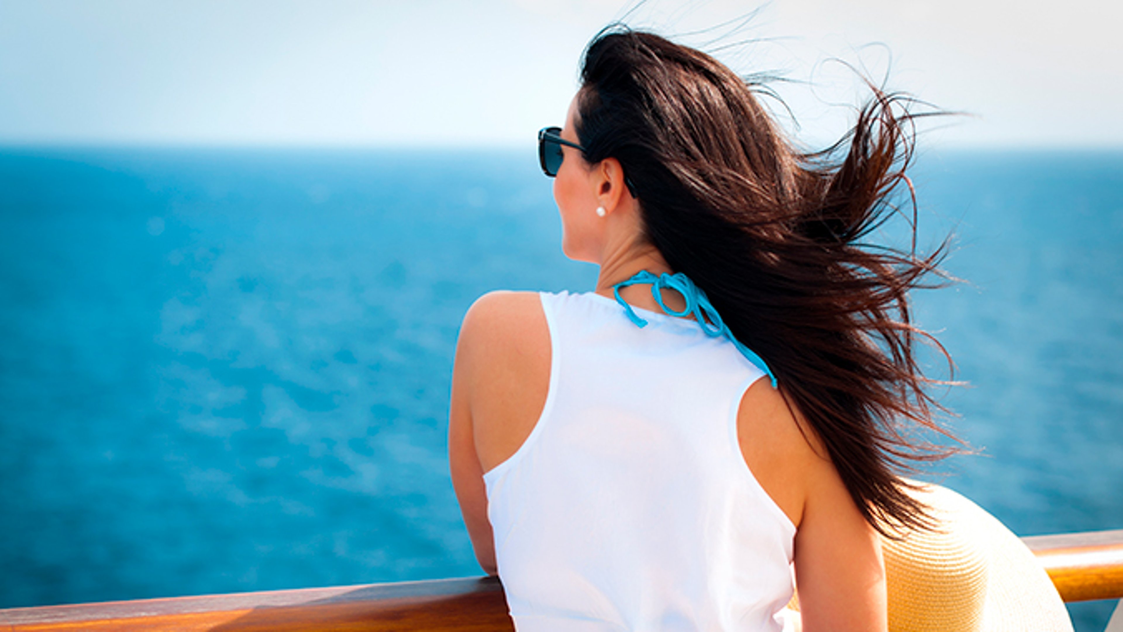 Woman holding straw hat with her back turned overlooking deck of cruise ship on blue ocean in background