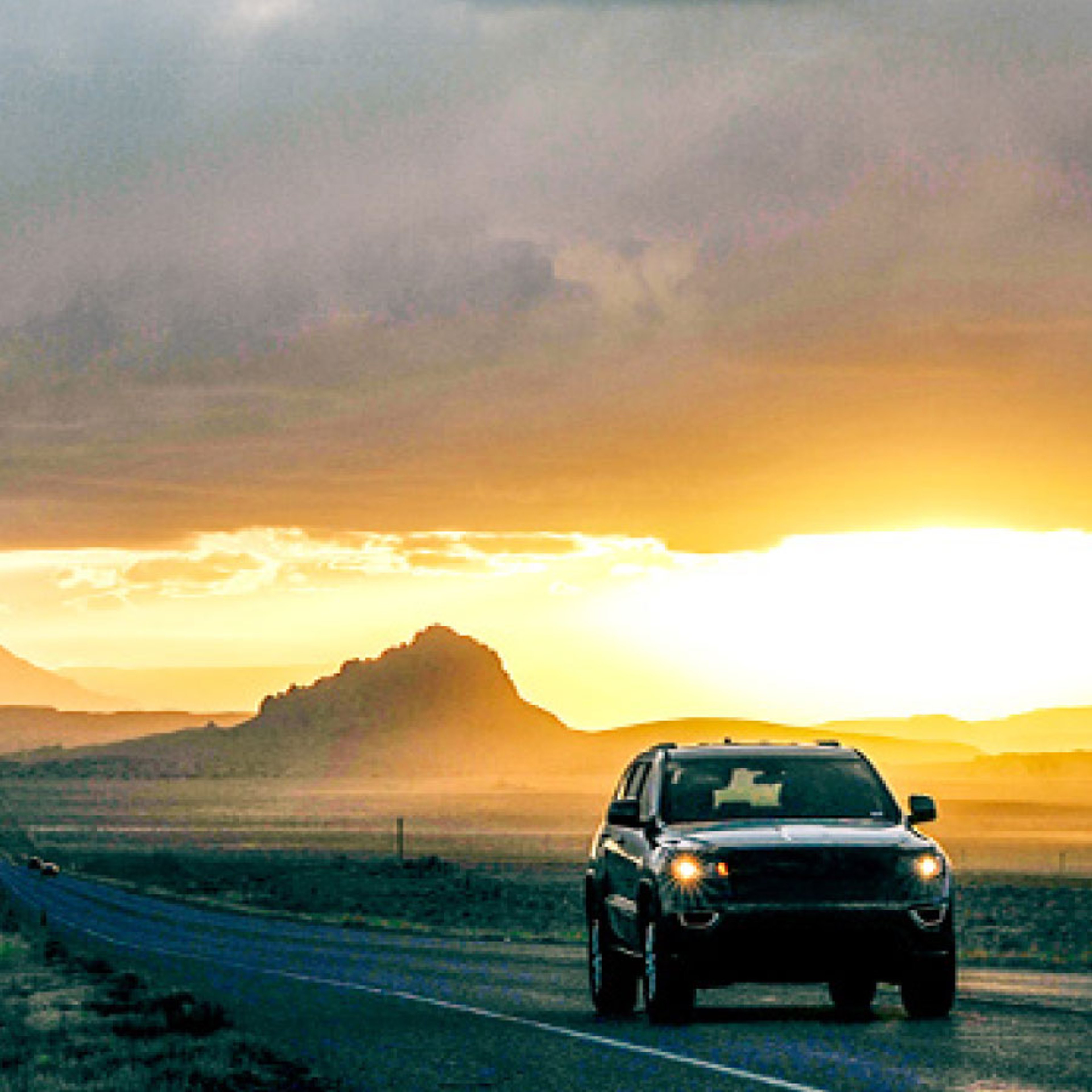 SUV driving along a desert highway at sunset, with mountains and dramatic golden clouds in the background.