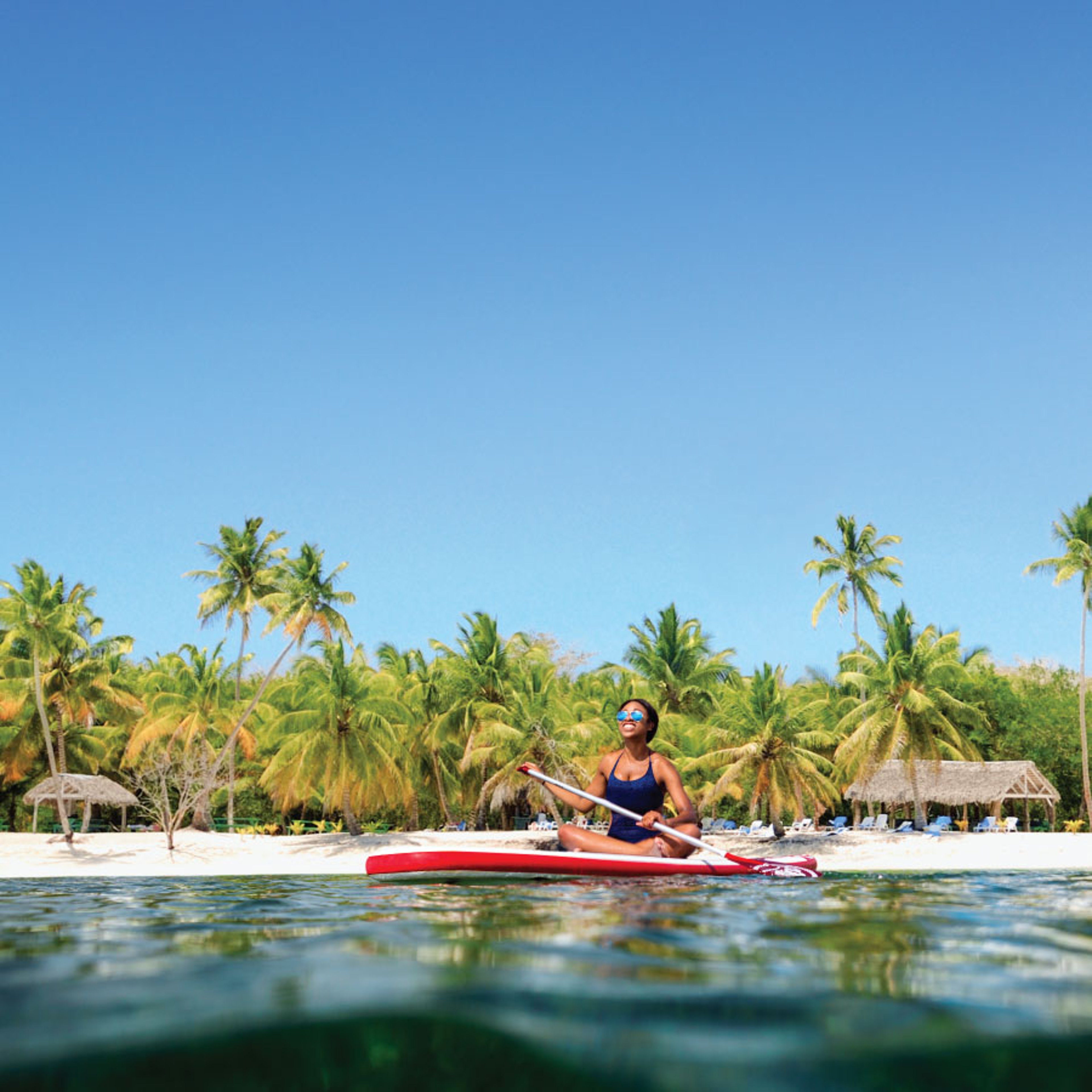 Woman sitting on a paddle board in the ocean near a tropical shoreline, with palm trees and a white sandy beach visible in the background.