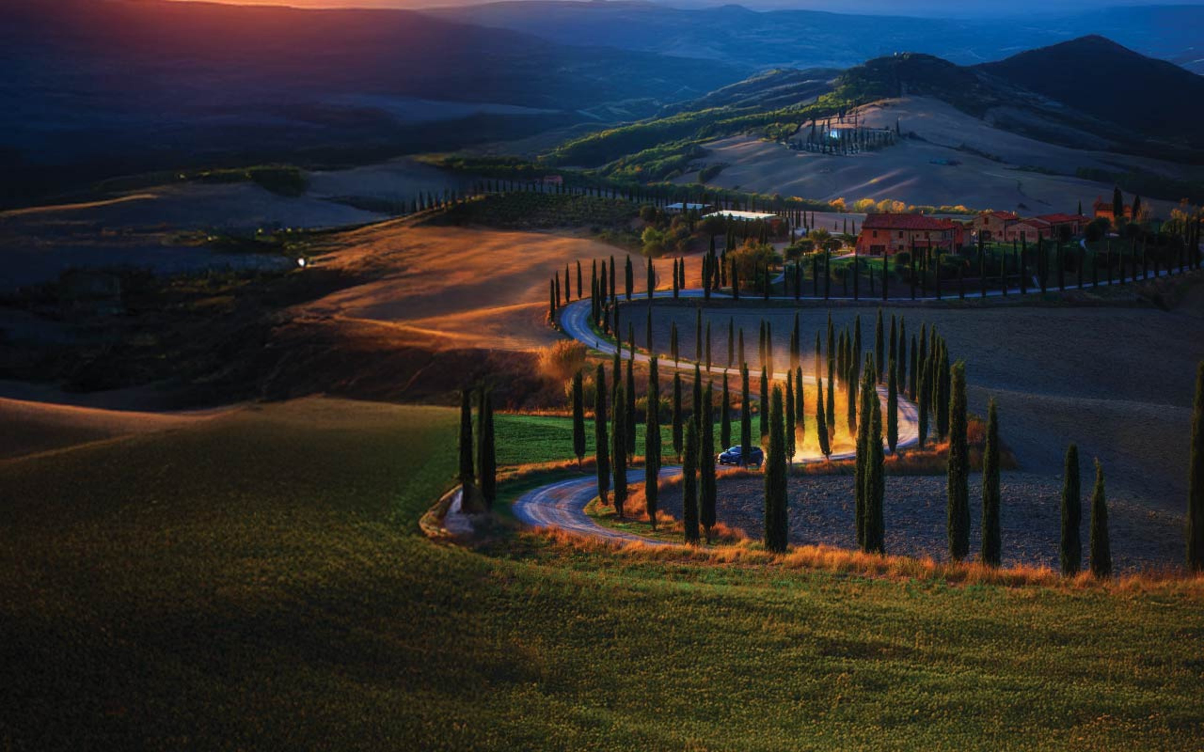 View of a sunset over a mountainside with scattered trees along a road with an SUV driving along it