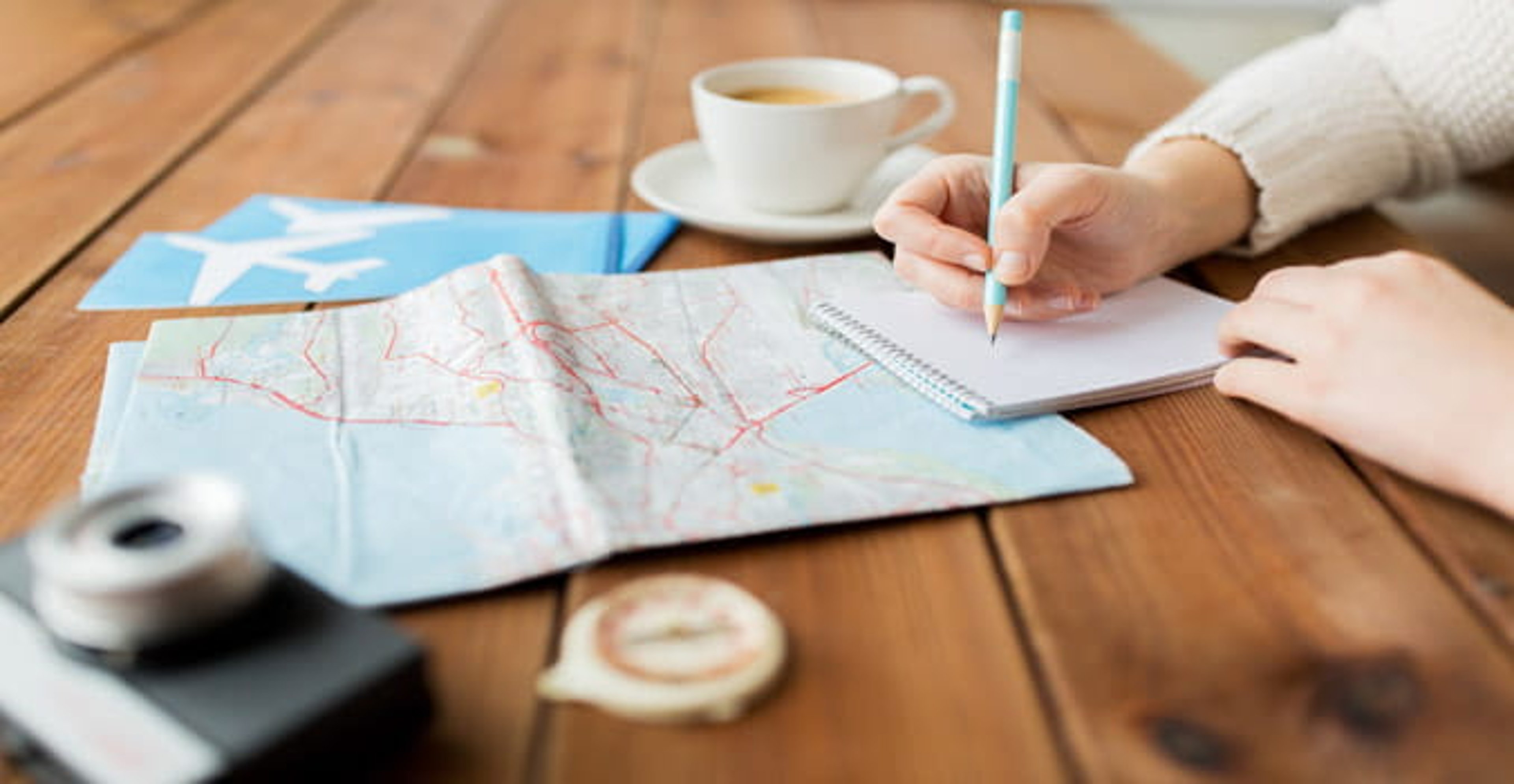 A person writing a note on a table surrounded by a map, documents, camera, and a cup of coffee