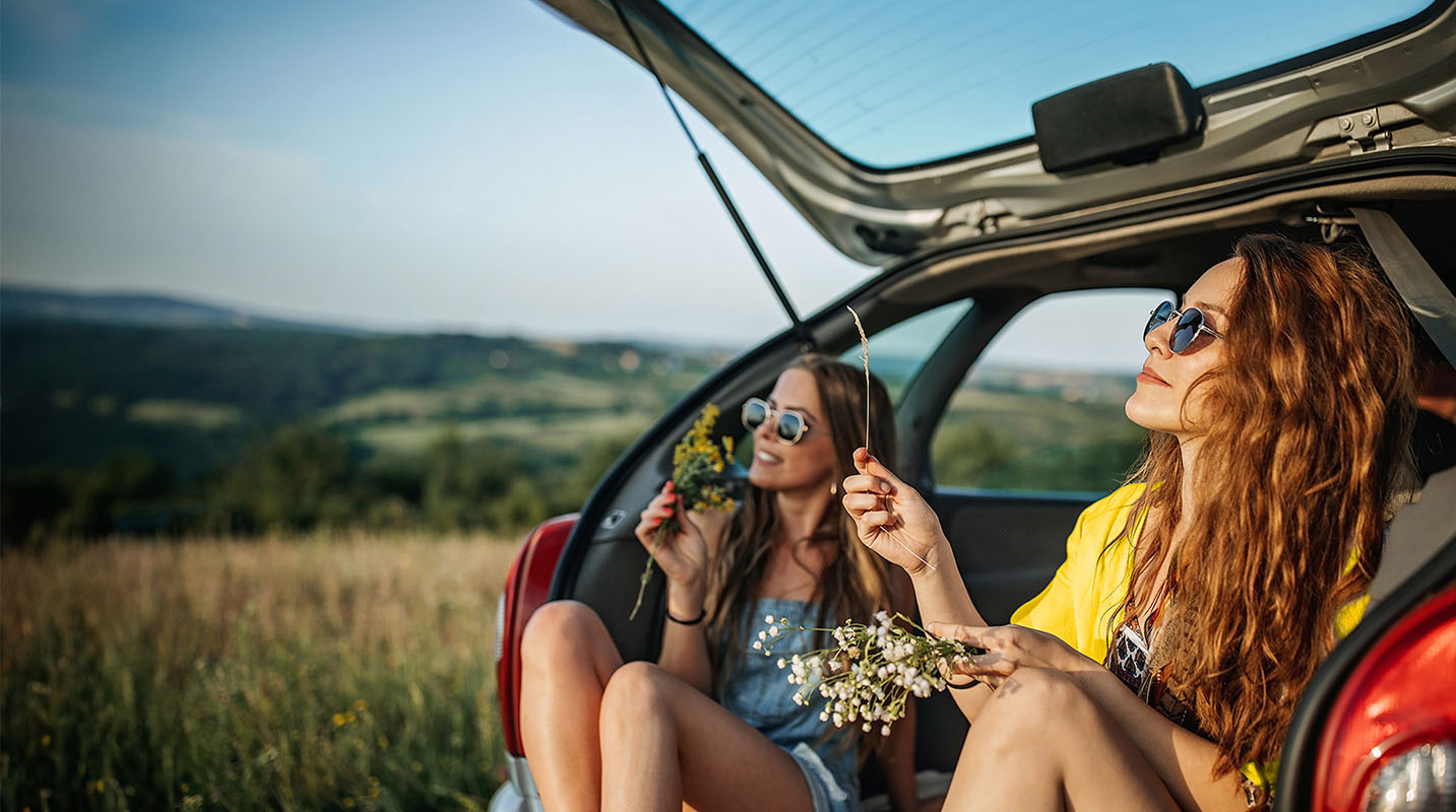 Two young women sitting in open trunk of rental car while on vacation with open grassland and mountains in the background