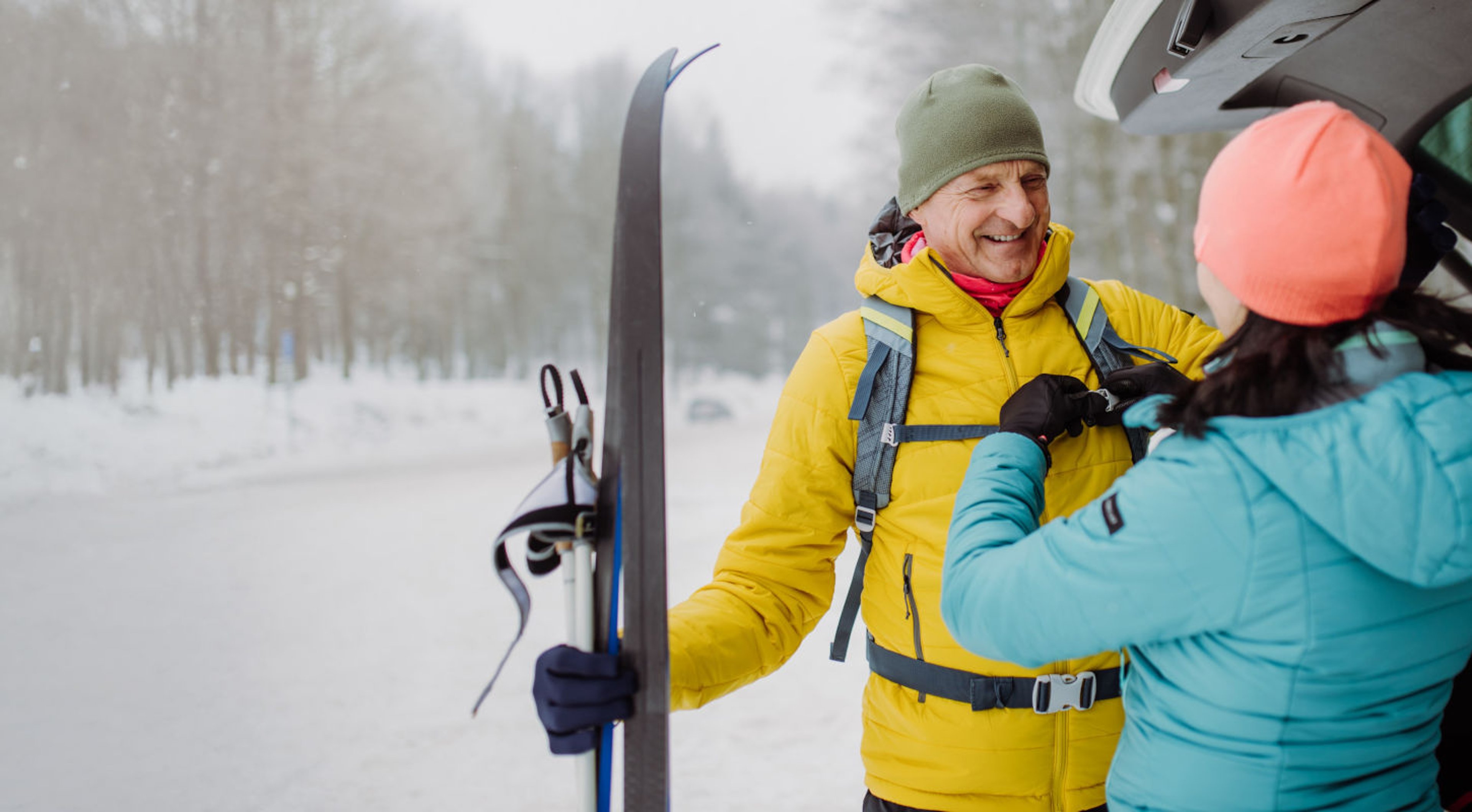 Smiling man with yellow jacket holding skis on snowy slope while talking to woman in ski gear