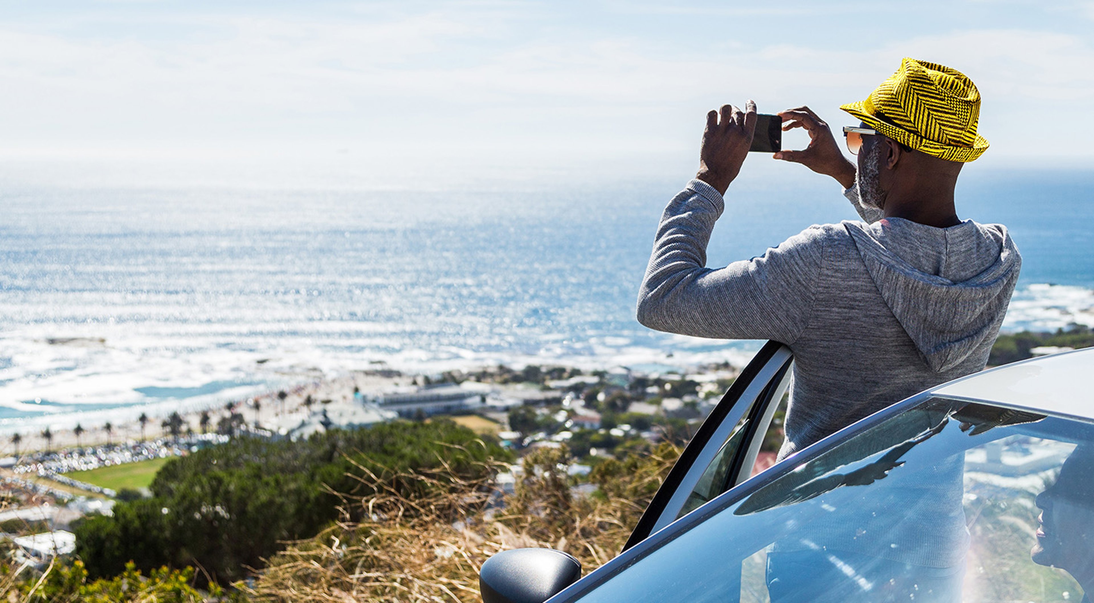 Man in yellow hat leaning against rental car taking picture of seaside coast while on vacation