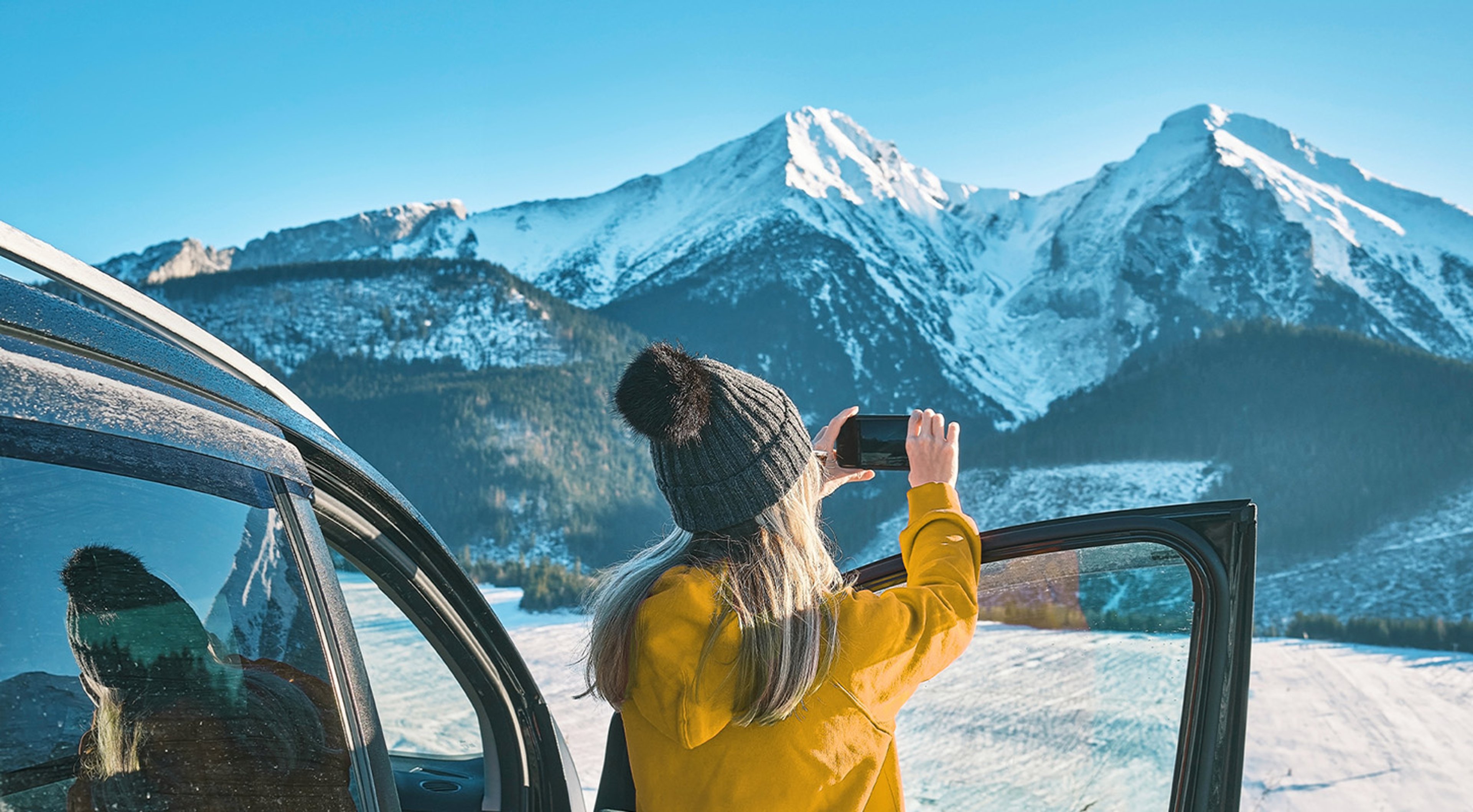 Woman with ski hat standing beside her car rental taking picture on her smartphone of snowy mountains