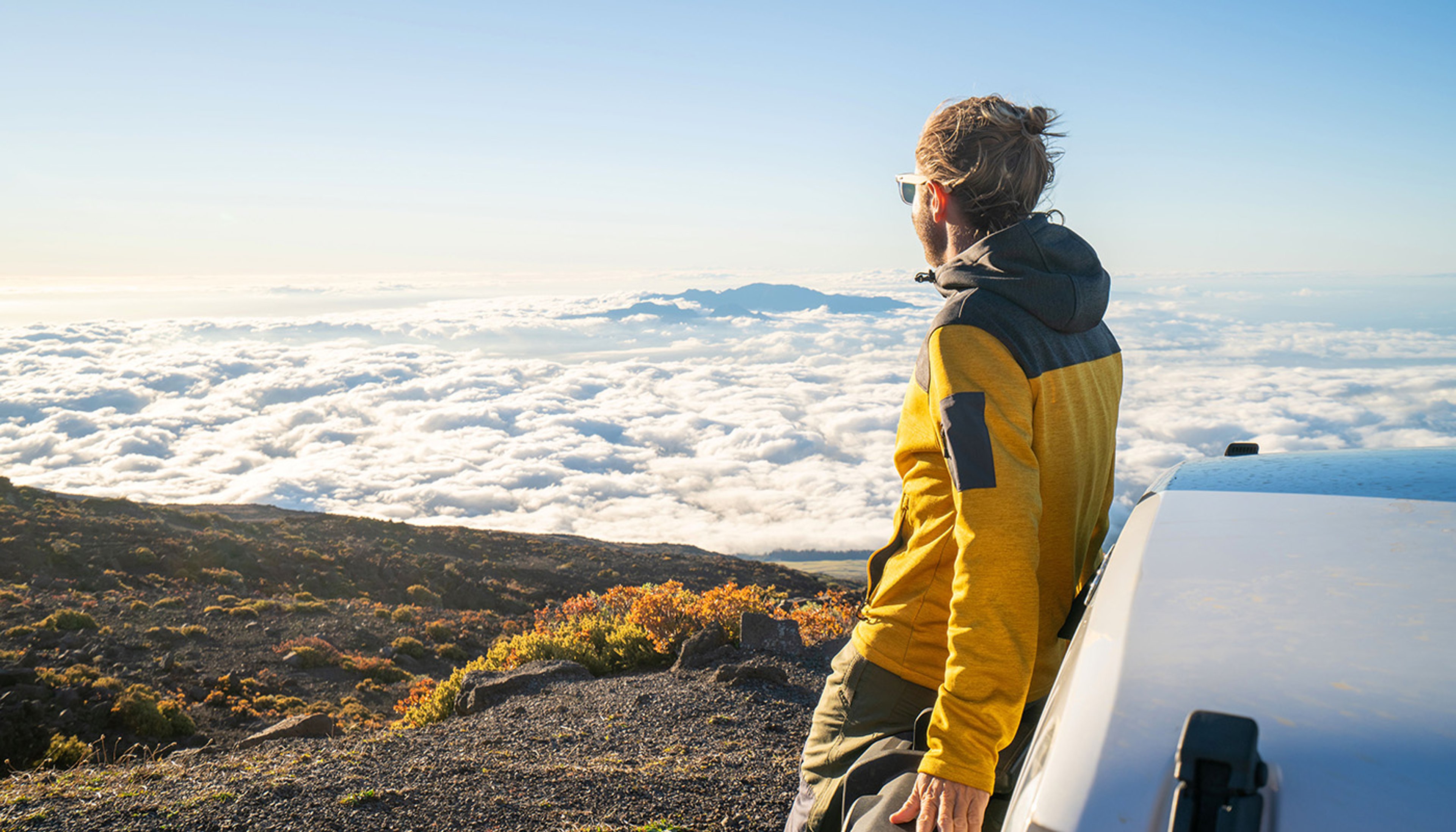 Man standing against rental car atop mountain with view of clouds below on sunny day