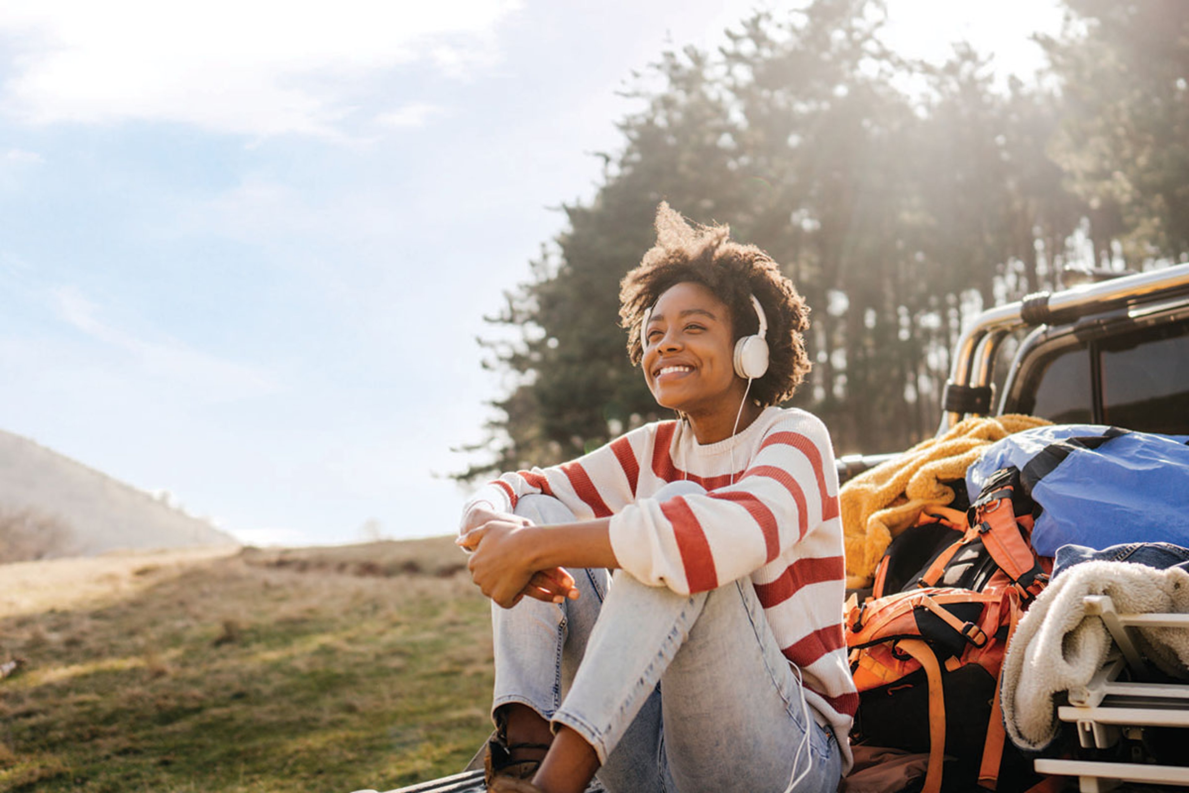 A smiling woman wearing headphones sits on the ground beside an open car trunk filled with bags and blankets, enjoying a sunny outdoor moment near a wooded area.