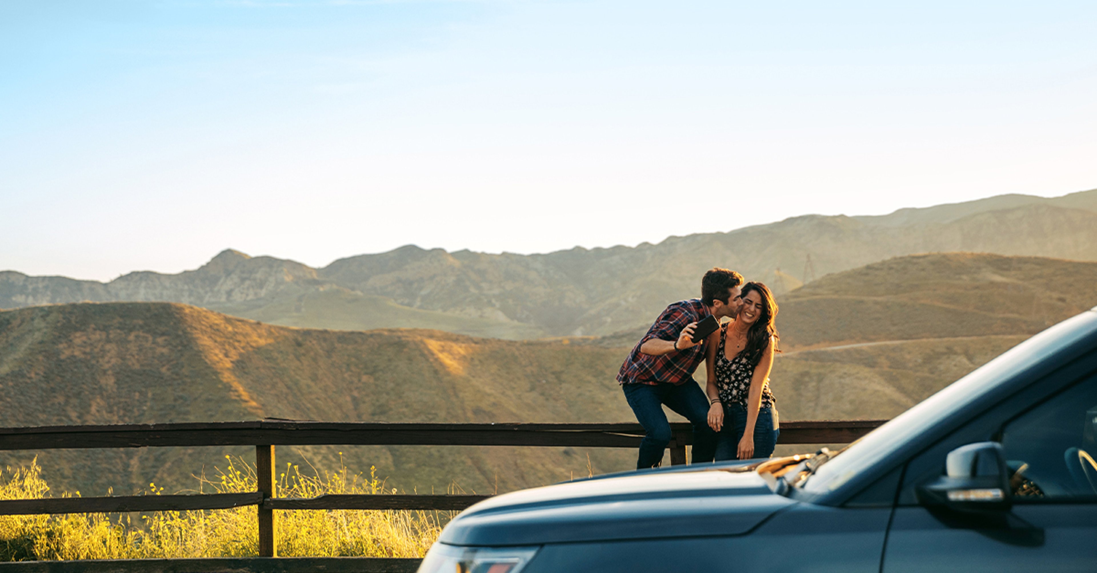 A couple sits on a wooden fence with mountains in the background, while a car is parked nearby.