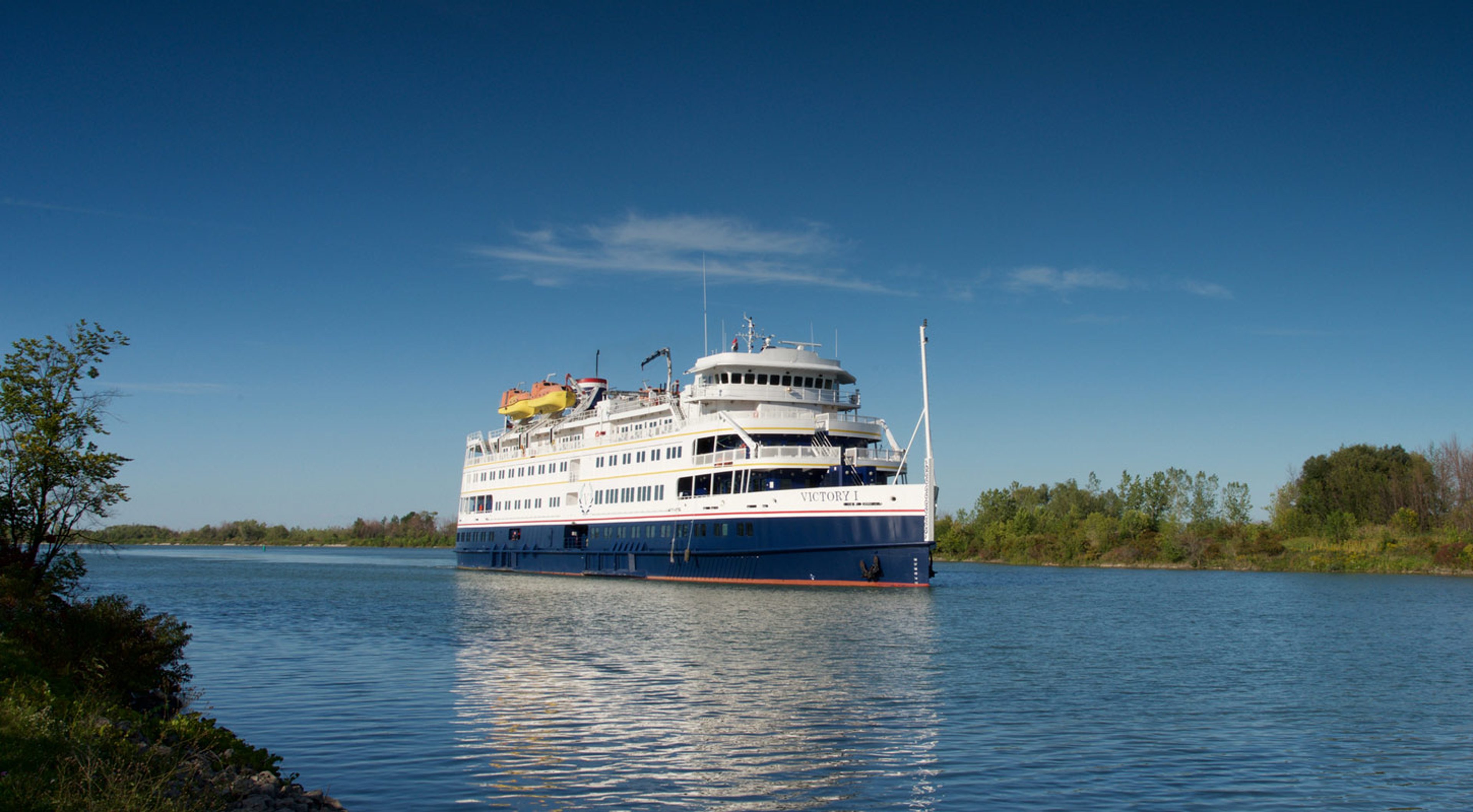 Victory Cruise Lines Victory 1 sailing on a calm river with a green shoreline and a clear blue sky
