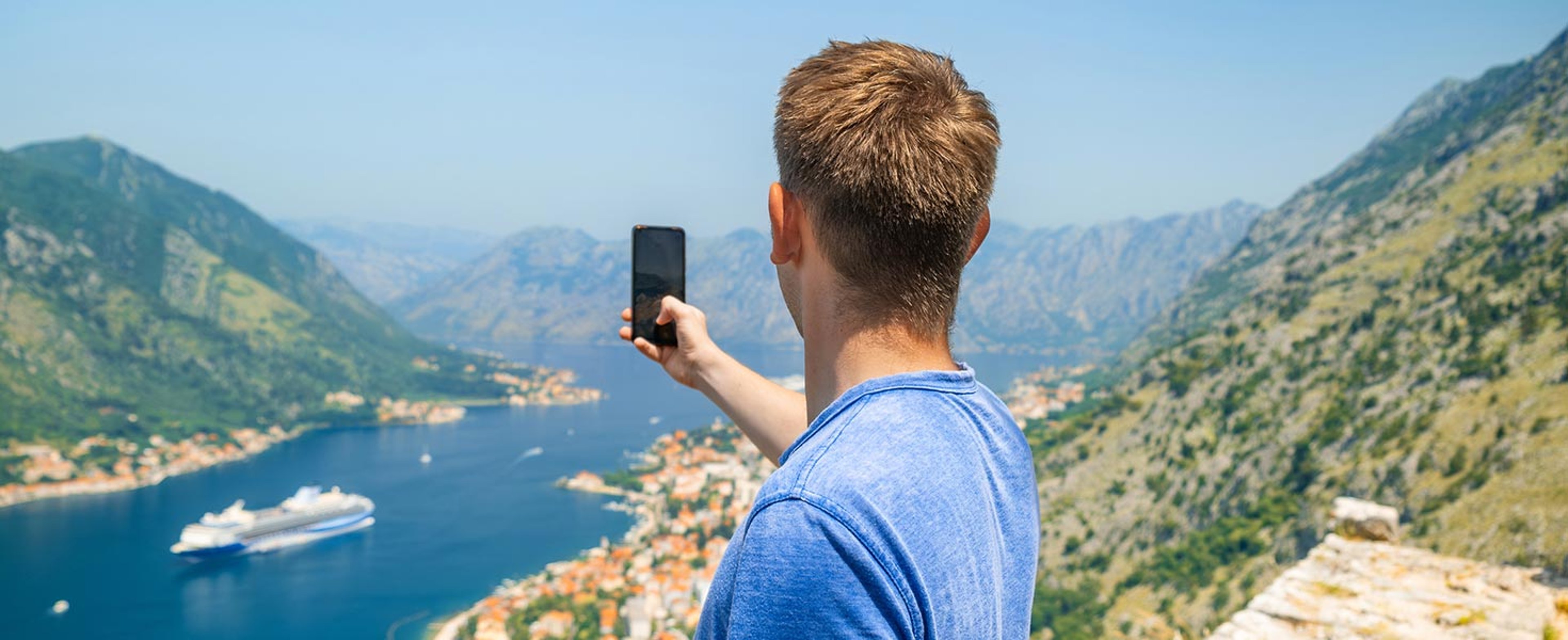 Young man taking image on phone of cruise ship in bay below 