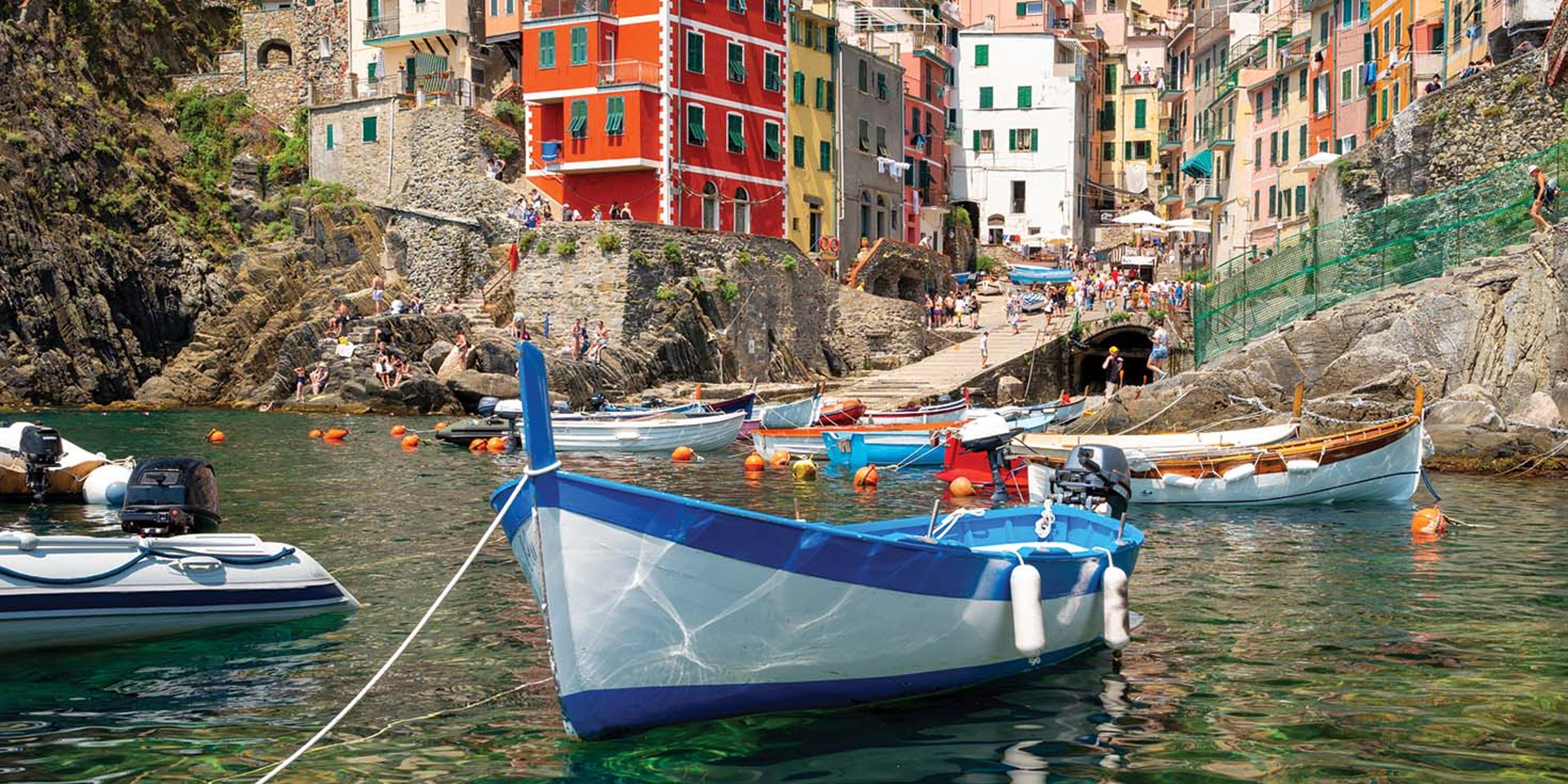 A harbor view of the Italian seaside village of Riomaggiore in Cinque Terre, with colorful, multi-story buildings built into the steep cliffs and small fishing boats docked in the turquoise water.