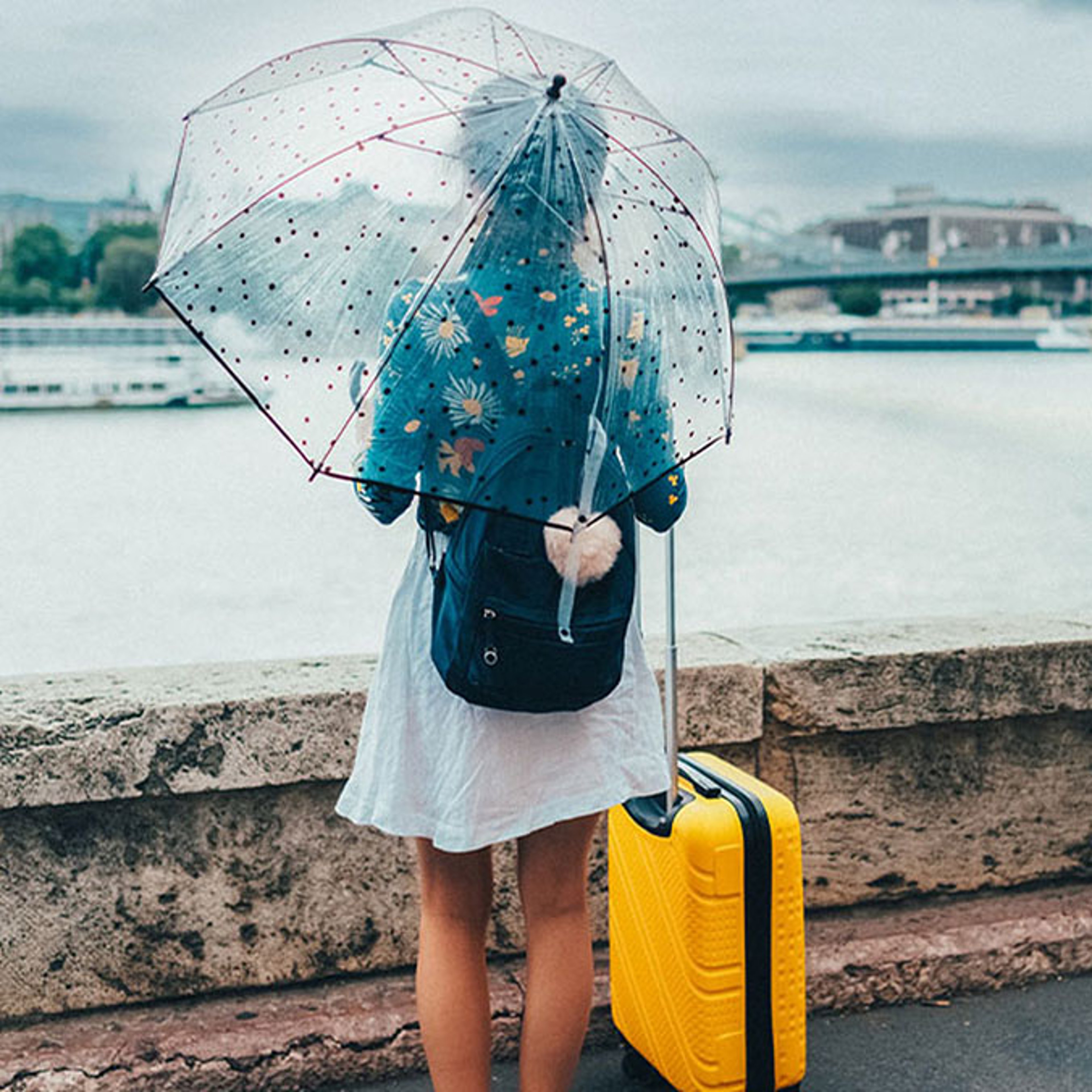 A woman with a clear umbrella stands in the rain with her yellow luggage, overlooking a scenic city view.