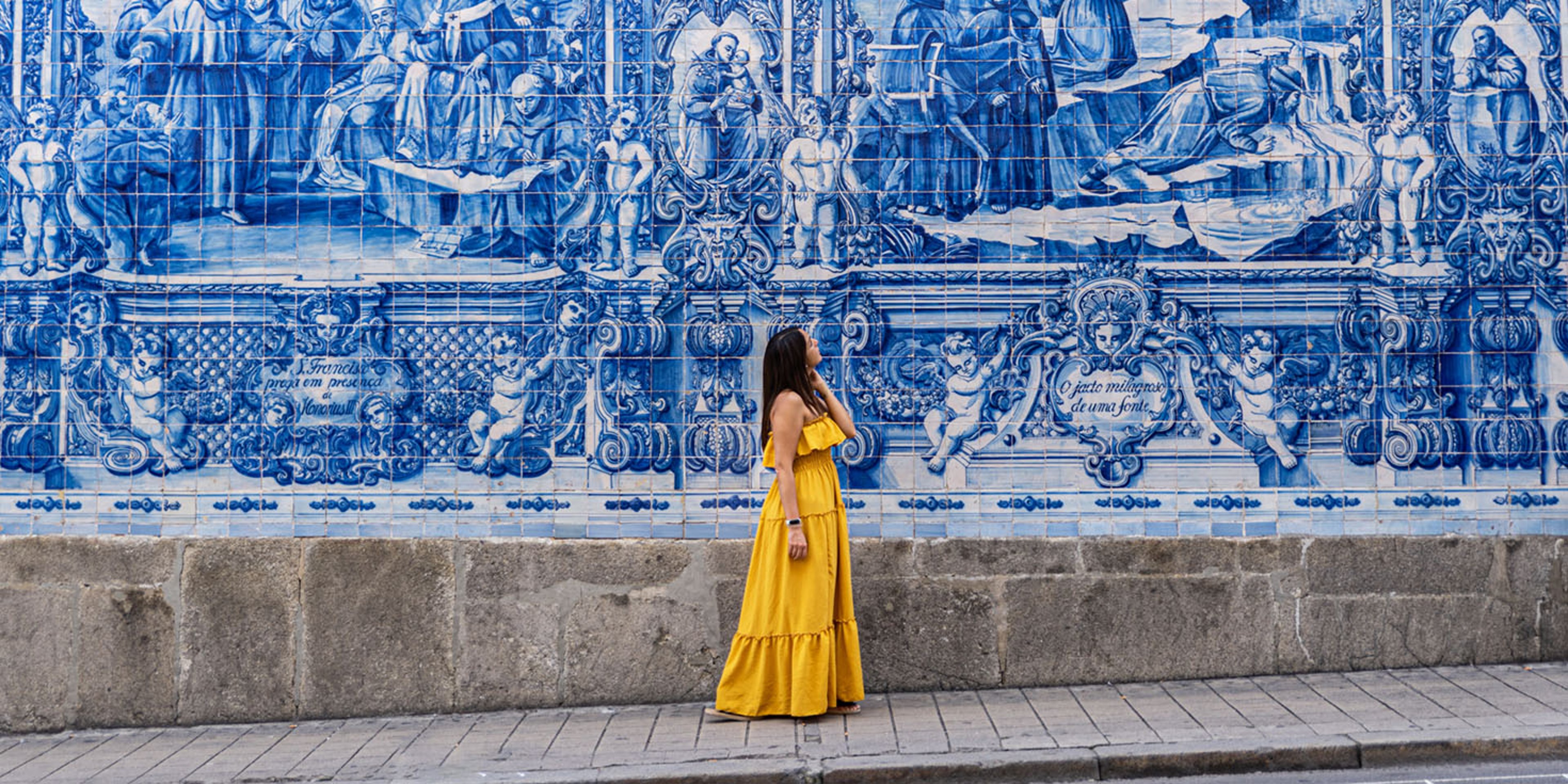 Woman in a yellow dress admiring the blue and white tiled facade of the Chapel of Souls in Porto, Portugal.