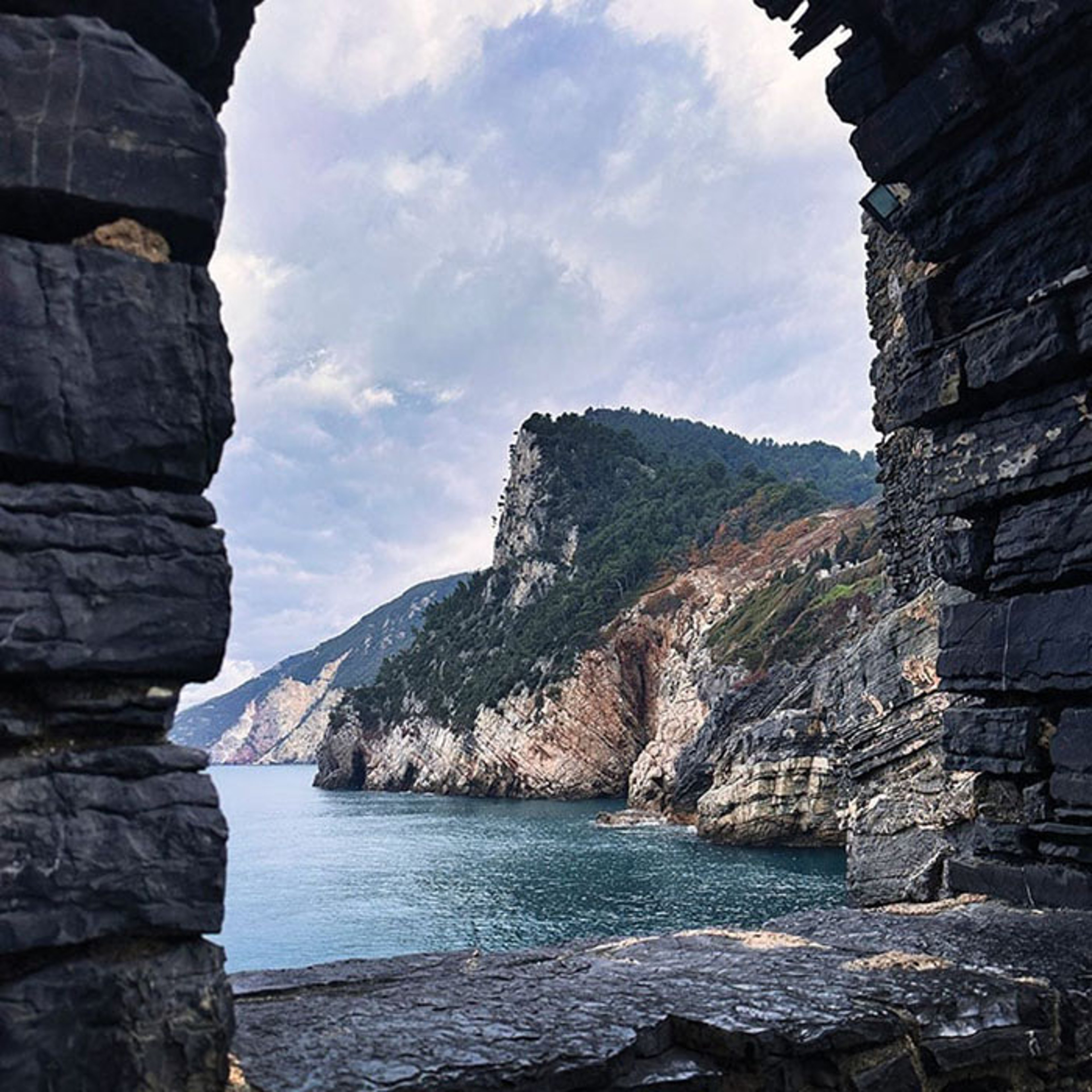 A scenic view of the Ligurian Sea and dramatic cliffs at Porto Venere, Italy, framed by a rugged stone archway under a cloudy sky.