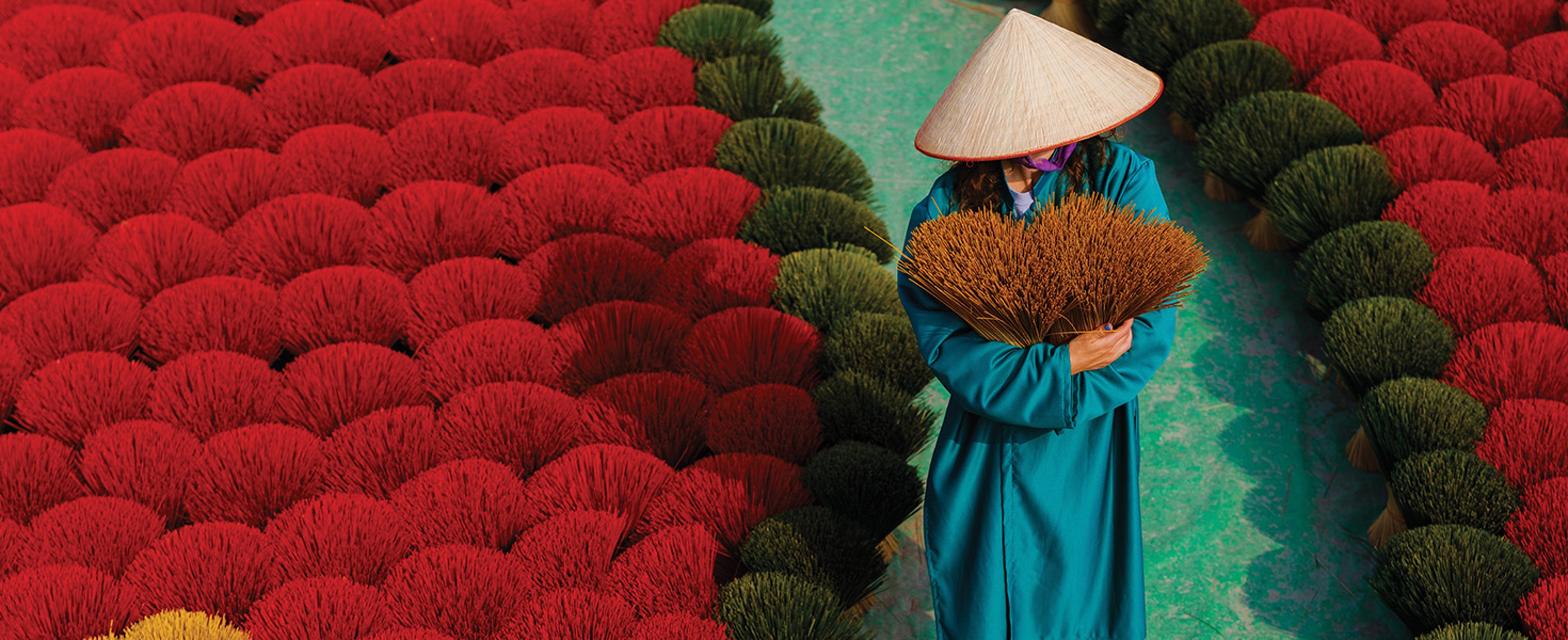 Woman in straw hat and holding bundles walking barefoot down a path in Hanoi Vietnam