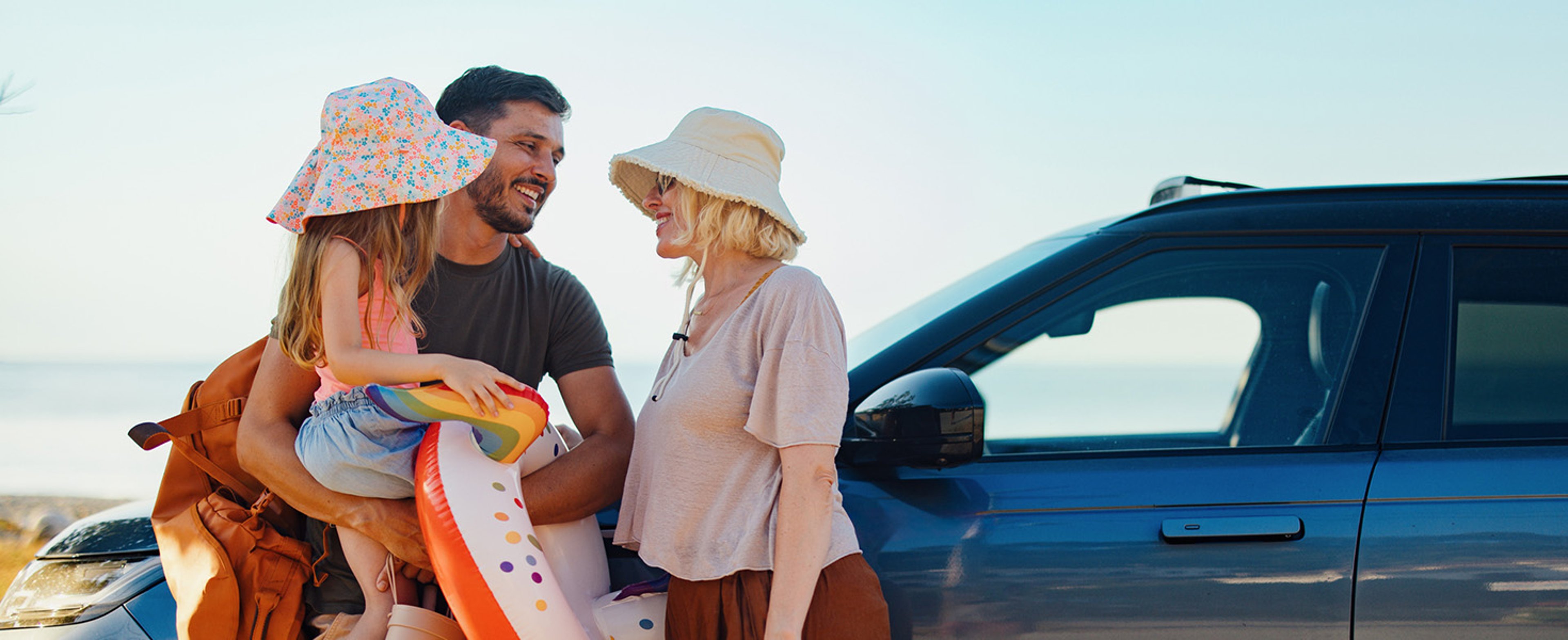 A smiling family of three standing next to a blue SUV on a sunny day, preparing for a beach trip with inflatable toys and a backpack.