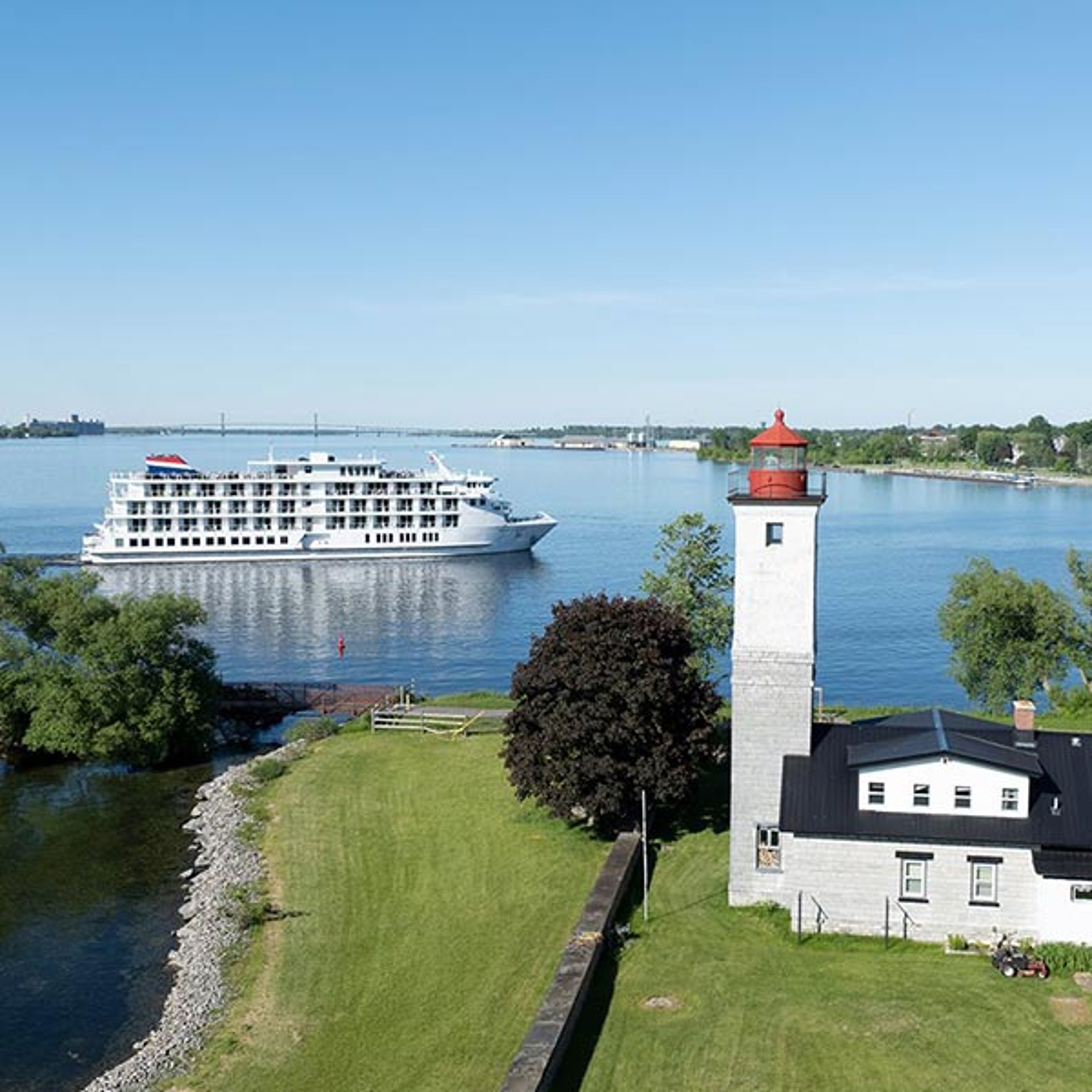 The image appears to show the Ogdensburg Harbor Lighthouse located at the confluence of the Oswegatchie and St. Lawrence rivers in Ogdensburg, New York, alongside a passing cruise ship that resembles the American Patriot from American Cruise Lines.