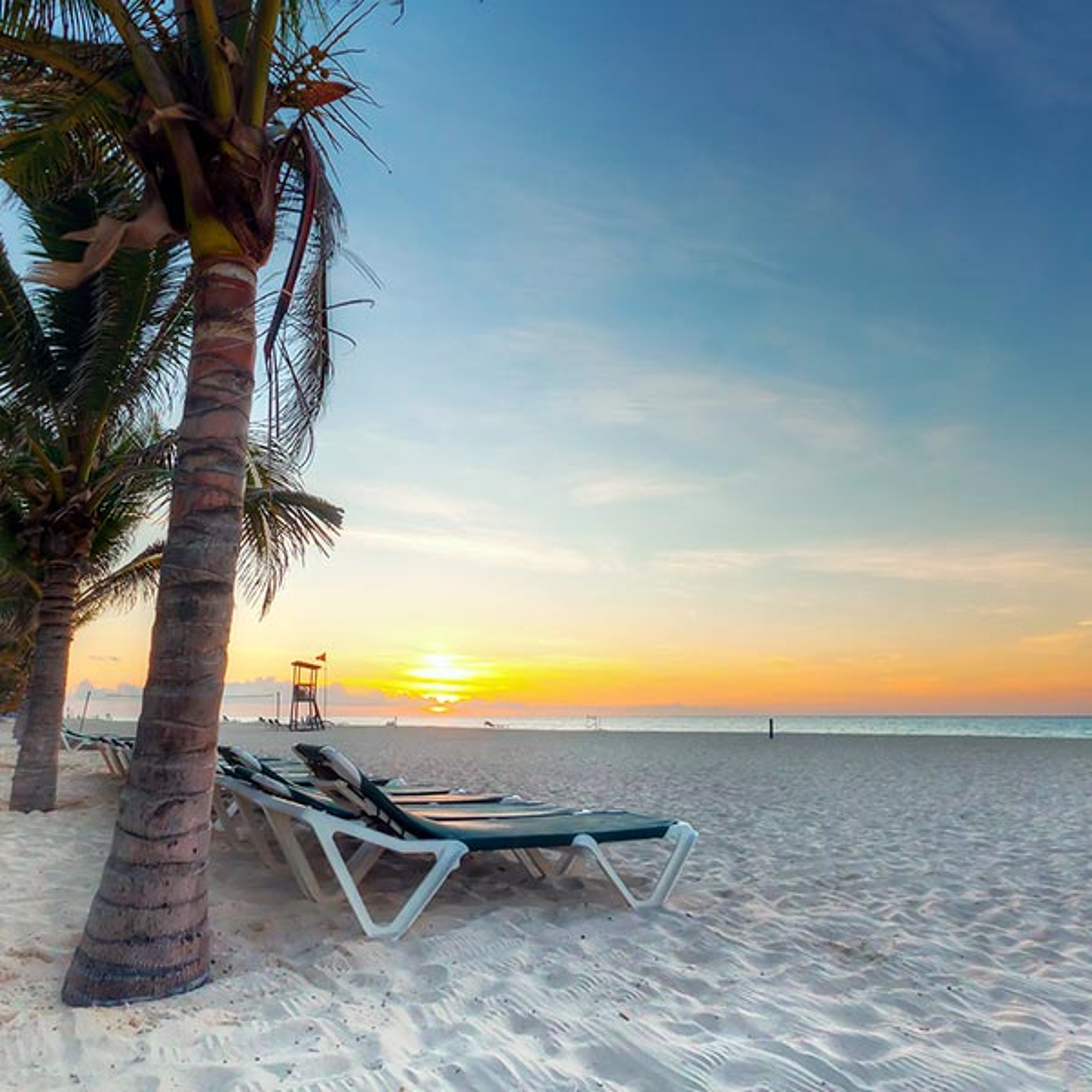 Lounge chair under palm trees on a Caribbean beach with sun setting over the water