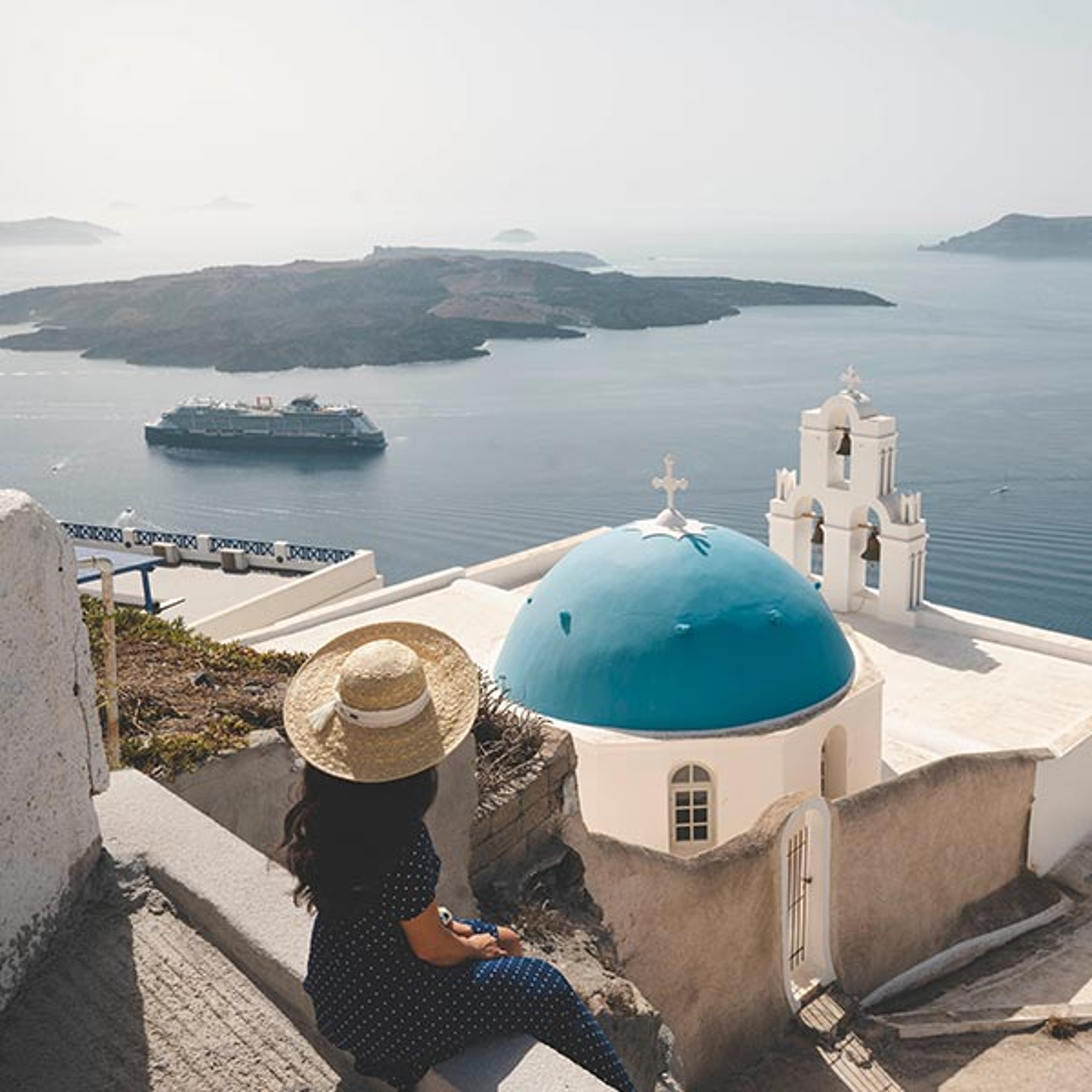 The image shows the iconic Three Bells of Fira blue-domed church overlooking the caldera in Santorini, Greece with a Celebrity ship in the background.