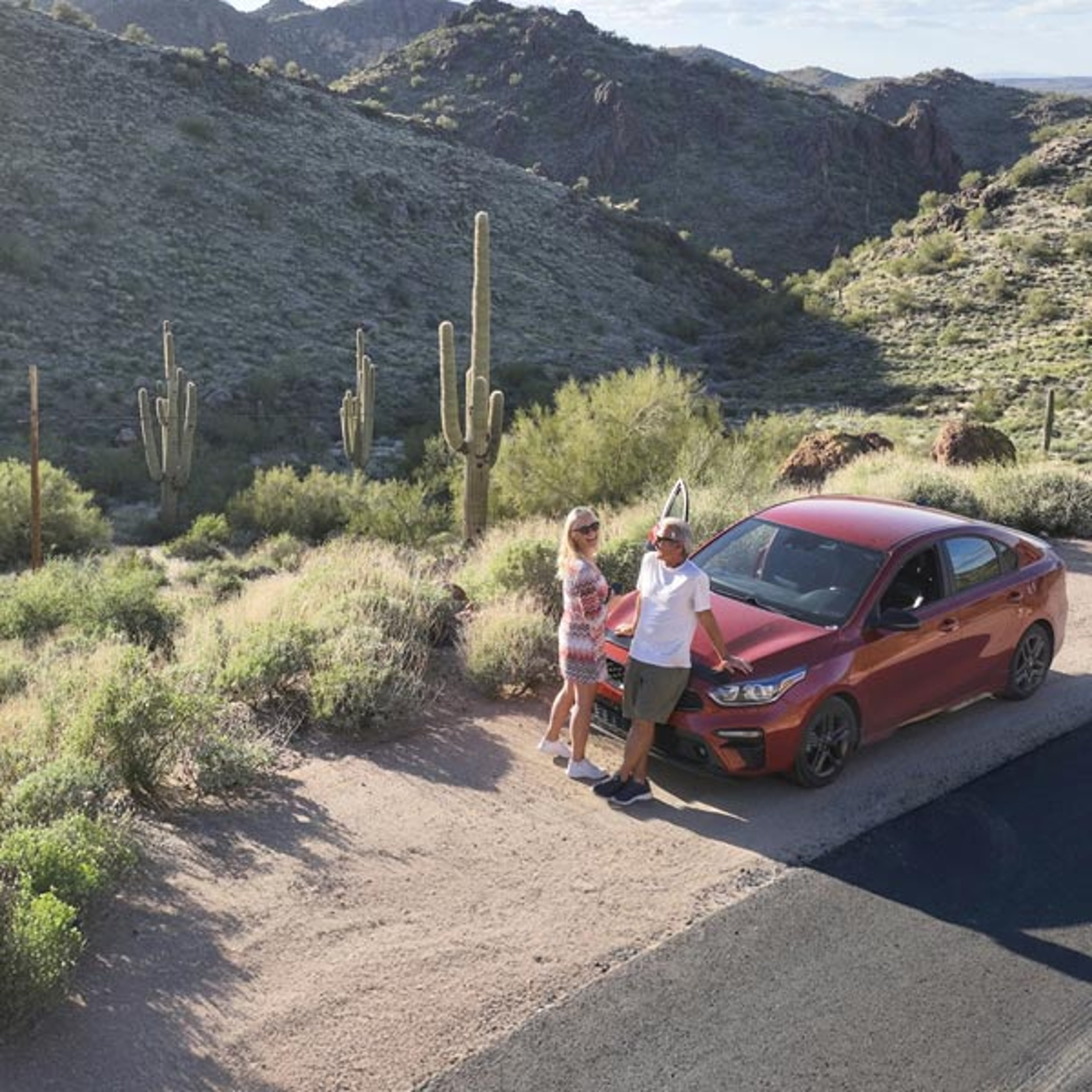 A couple leaning against the hood of their red car while parked along side the road in the dessert