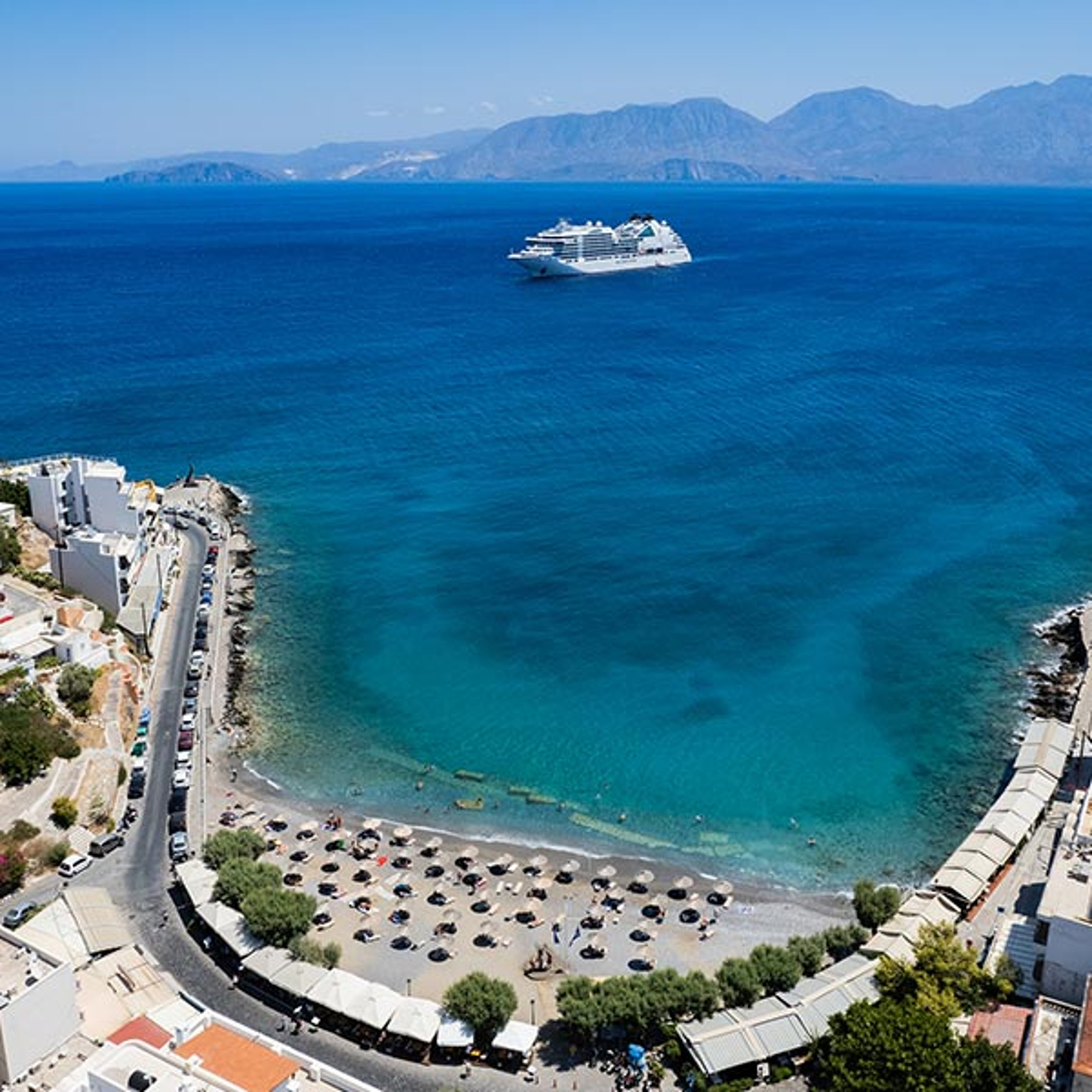 This image shows Kitroplateia Beach in the town of Agios Nikolaos on the island of Crete, Greece with a Seabourn ship docked in the background