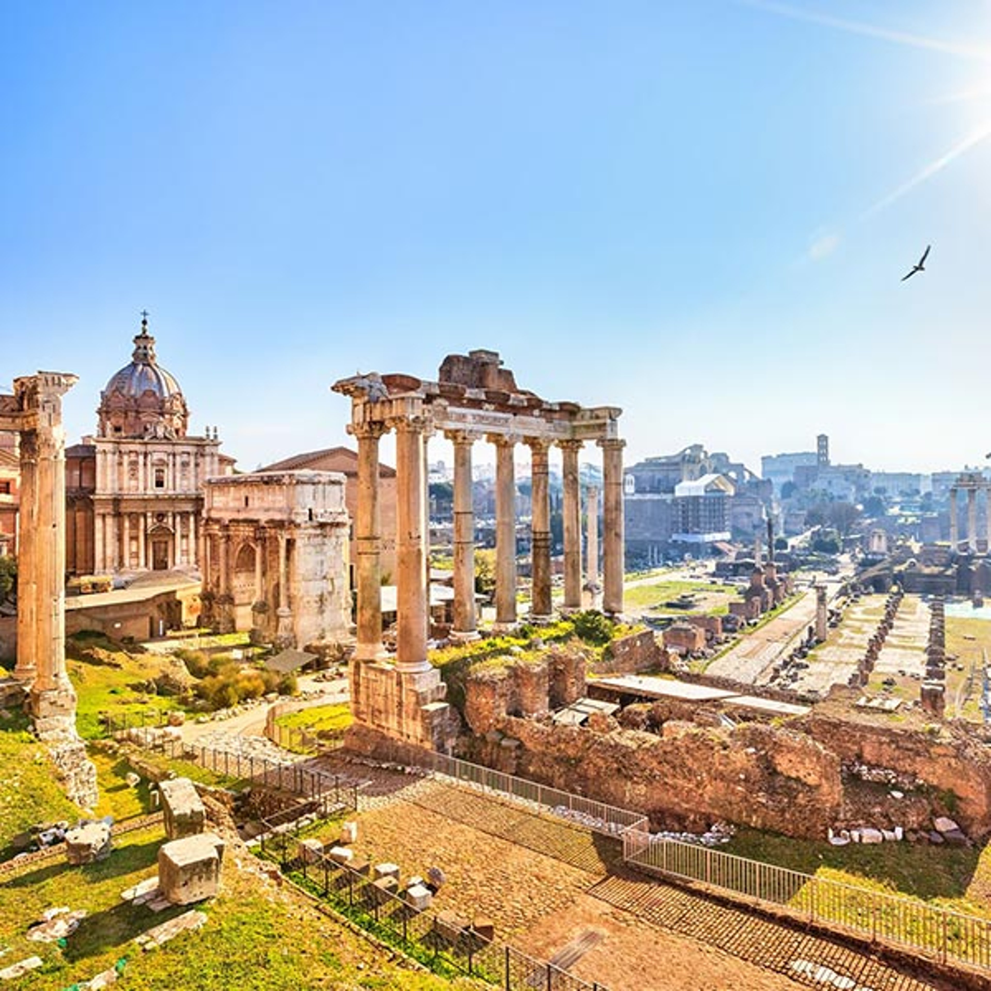 The image shows the ancient ruins of the Roman Forum in Rome, Italy, featuring weathered stone columns and remnants of classical architecture under a bright sky