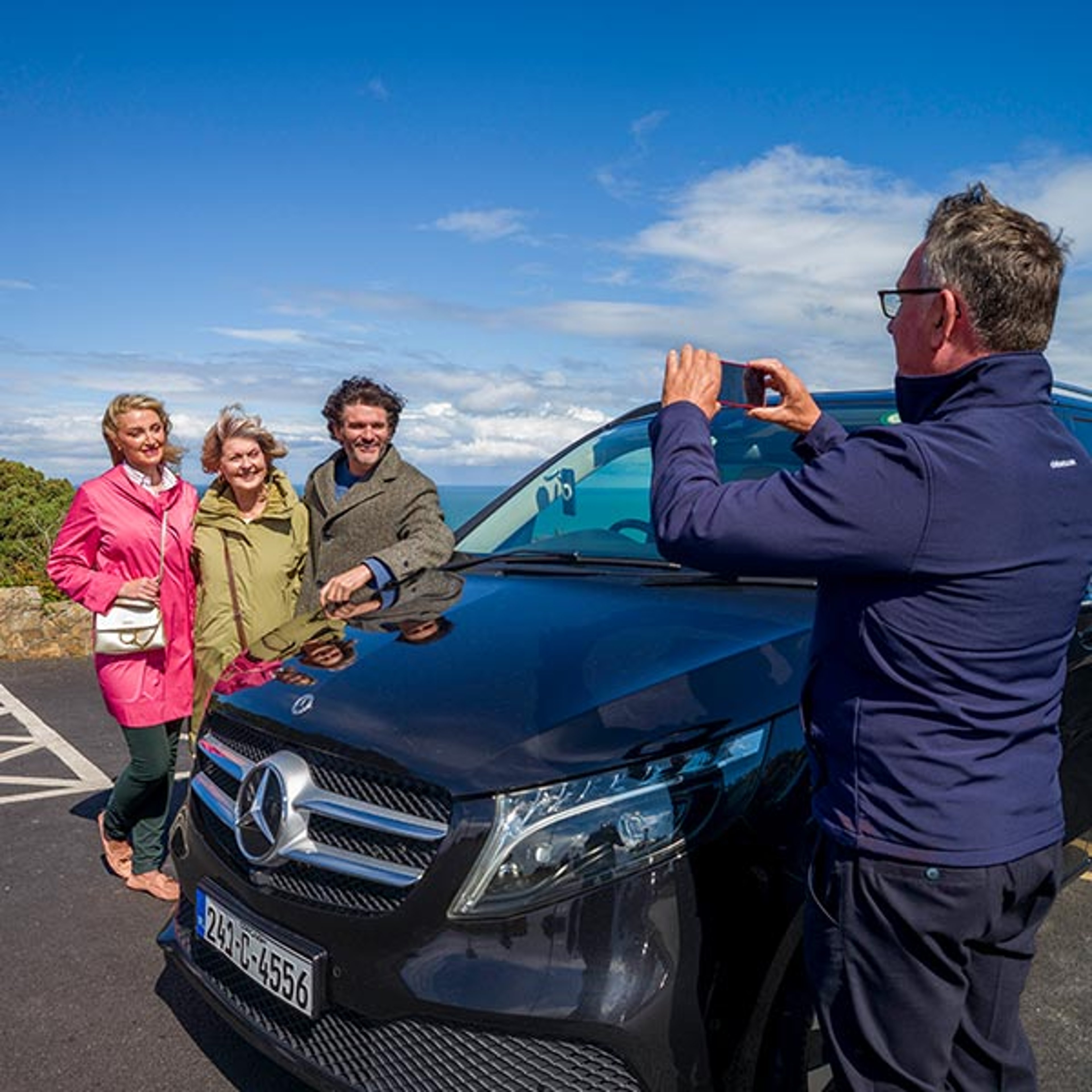 The image shows a group of tourists on a CIE Tours Private Driver journey posing in front of a dark Mercedes-Benz van while the driver takes their photo outdoors
