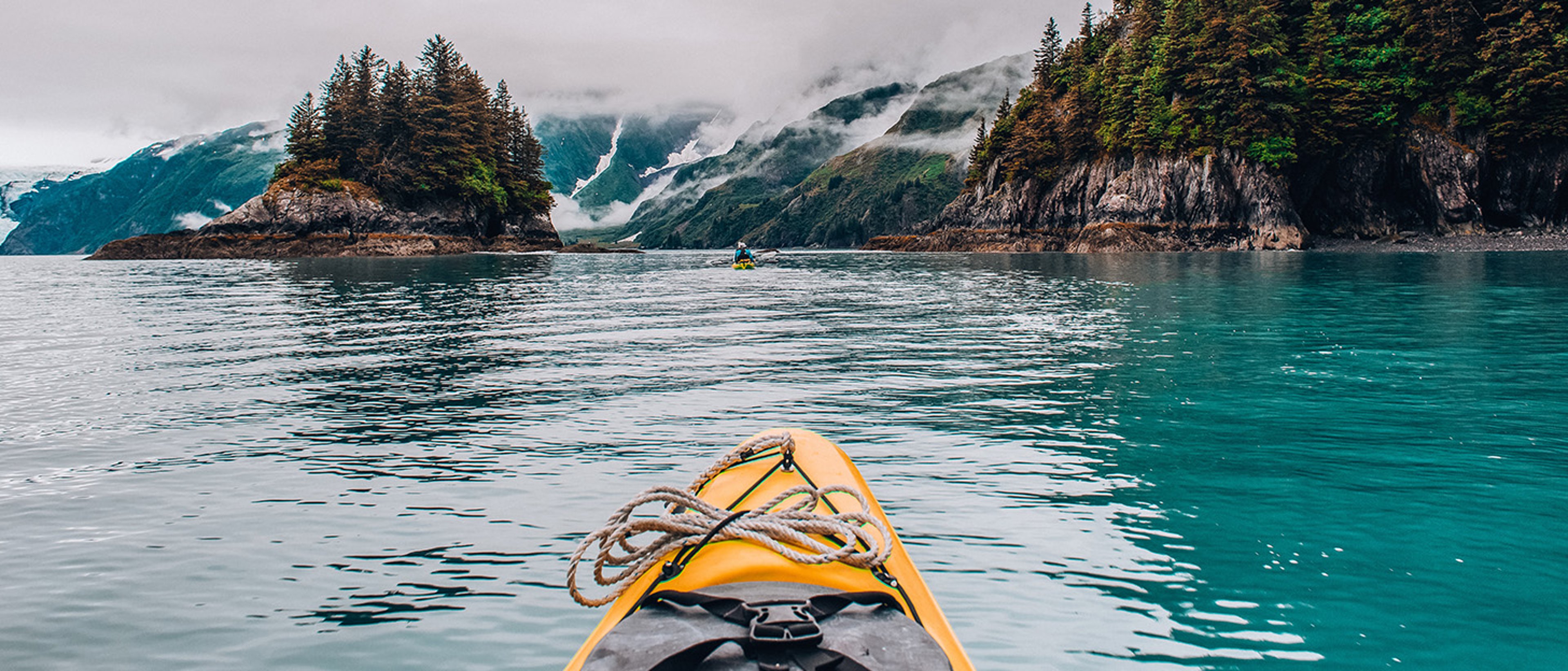 Yellow kayak with rope on its bow navigating through calm, turquoise waters surrounded by pine-covered rocky islands and misty mountains in the background.