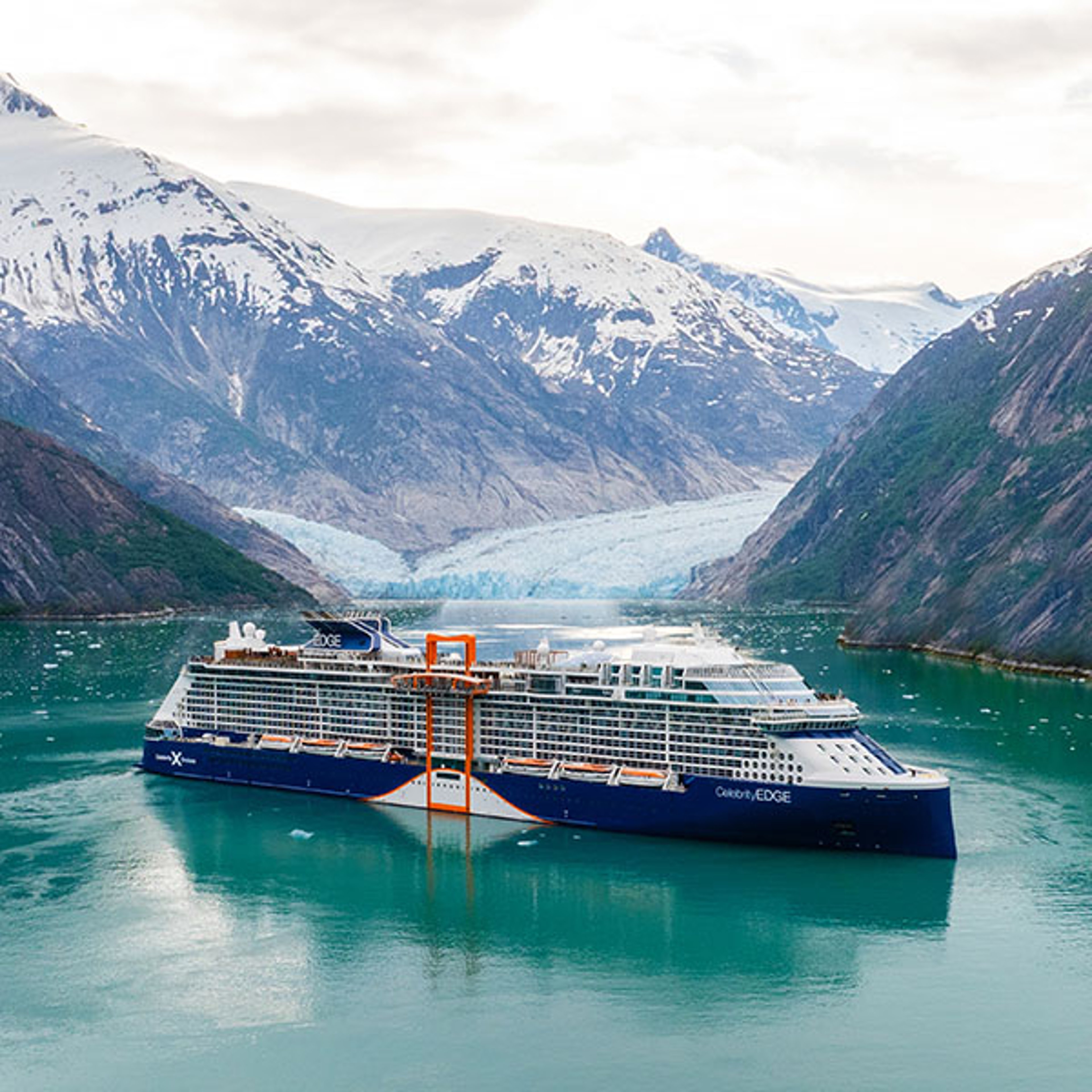 A large cruise ship sails on emerald green water in a glacial fjord, surrounded by snow-capped mountains and a distant glacier.