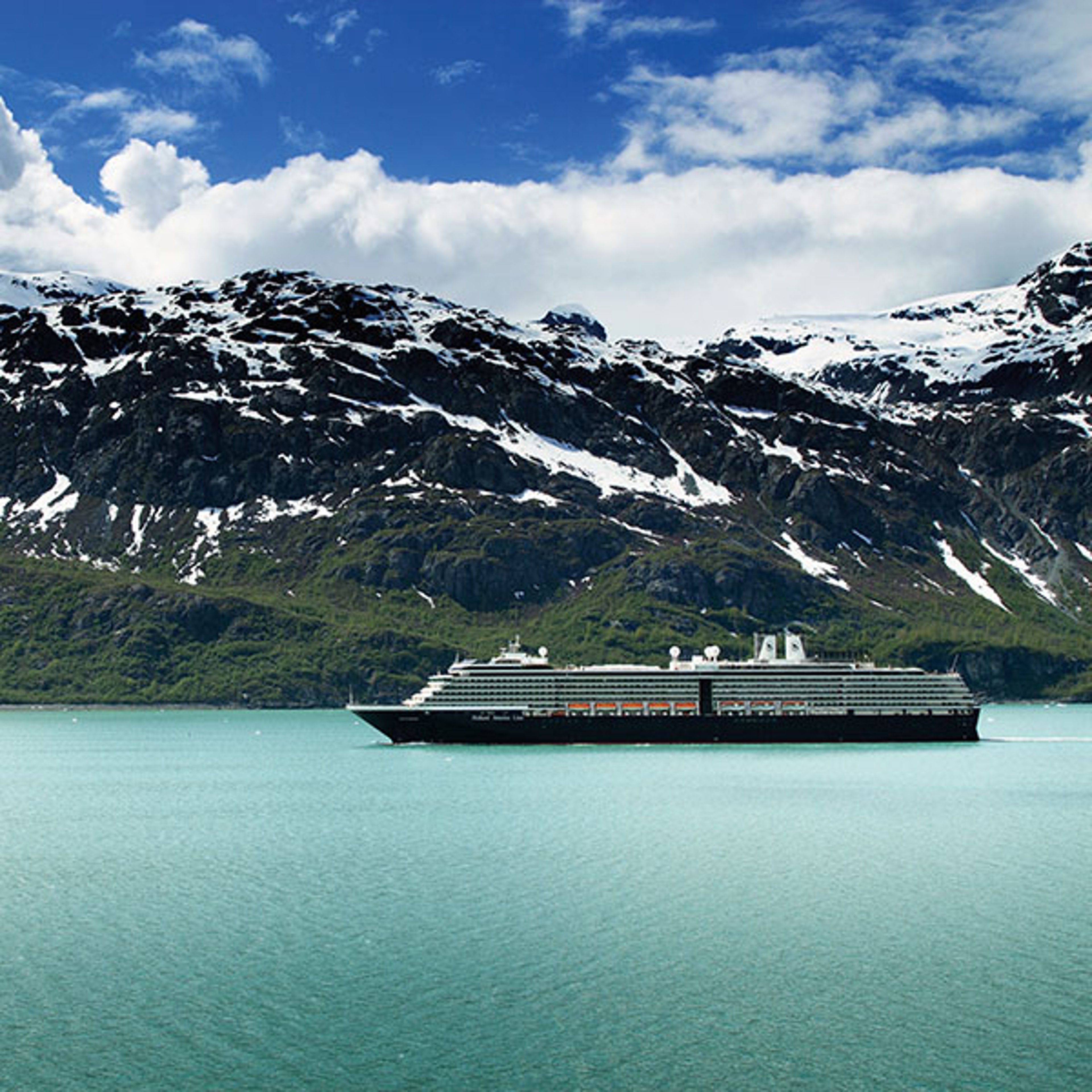 A large cruise ship sails through a narrow waterway surrounded by snow-capped mountains with a glacier visible in the background.