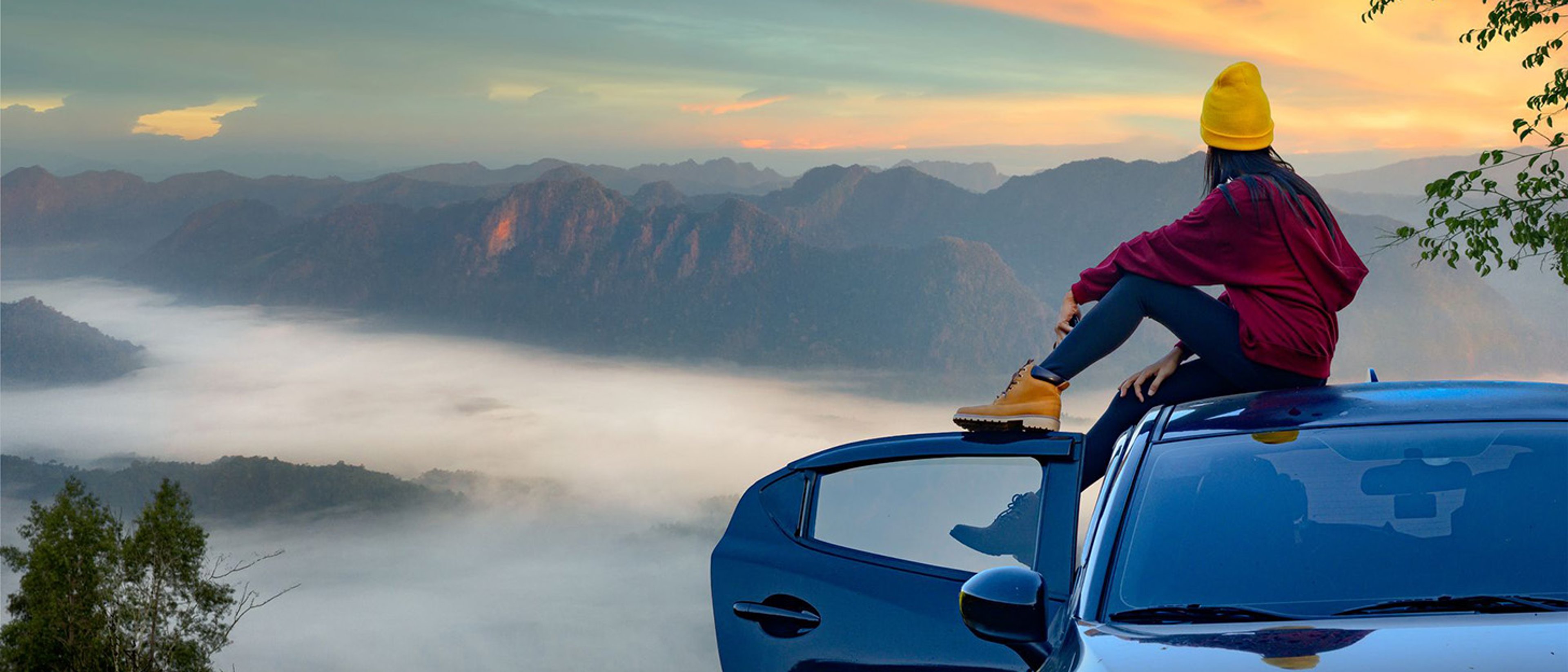 Woman with yellow hat and red jacket sits on top of car, overlooking scenic mountains