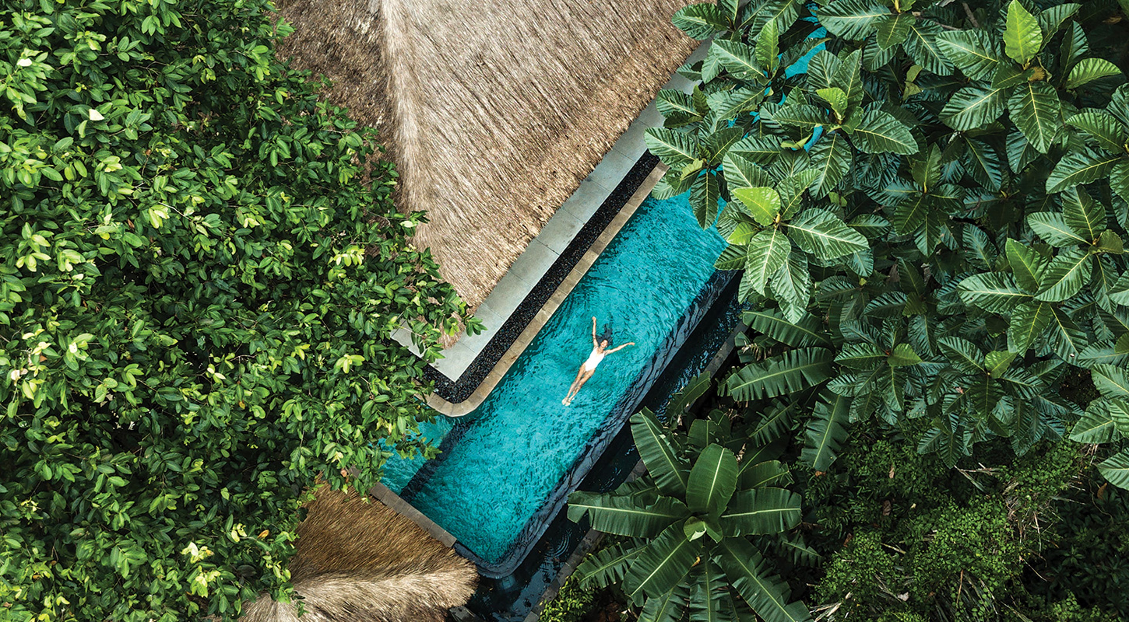 Aerial view of woman in white bathing suit floating in rectangular pool in rainforest on luxury travel vacation