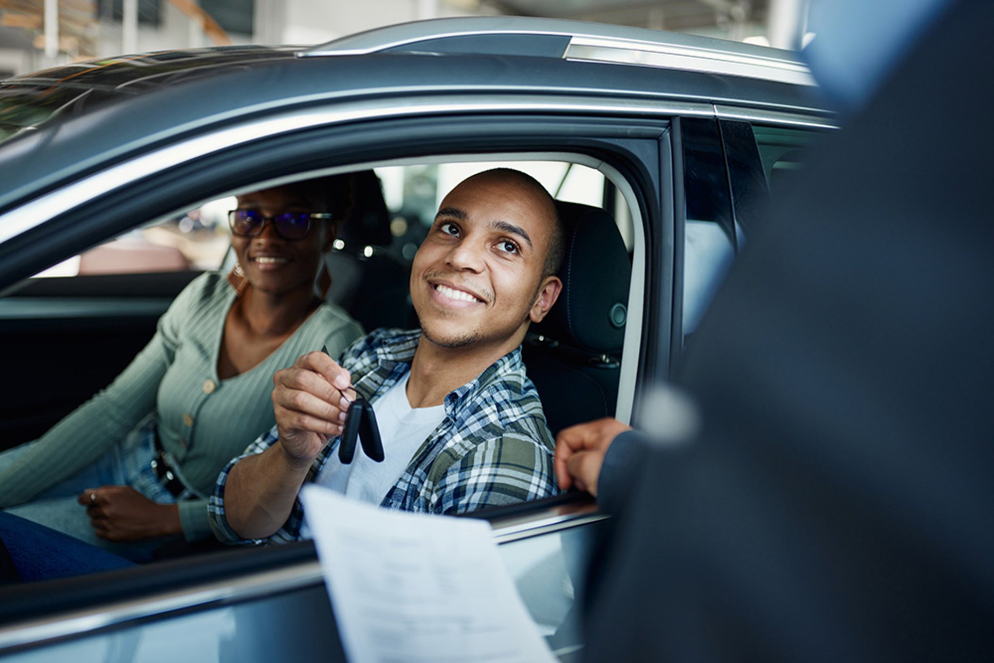 Smiling driver in car with female passenger taking keys from rental car representative