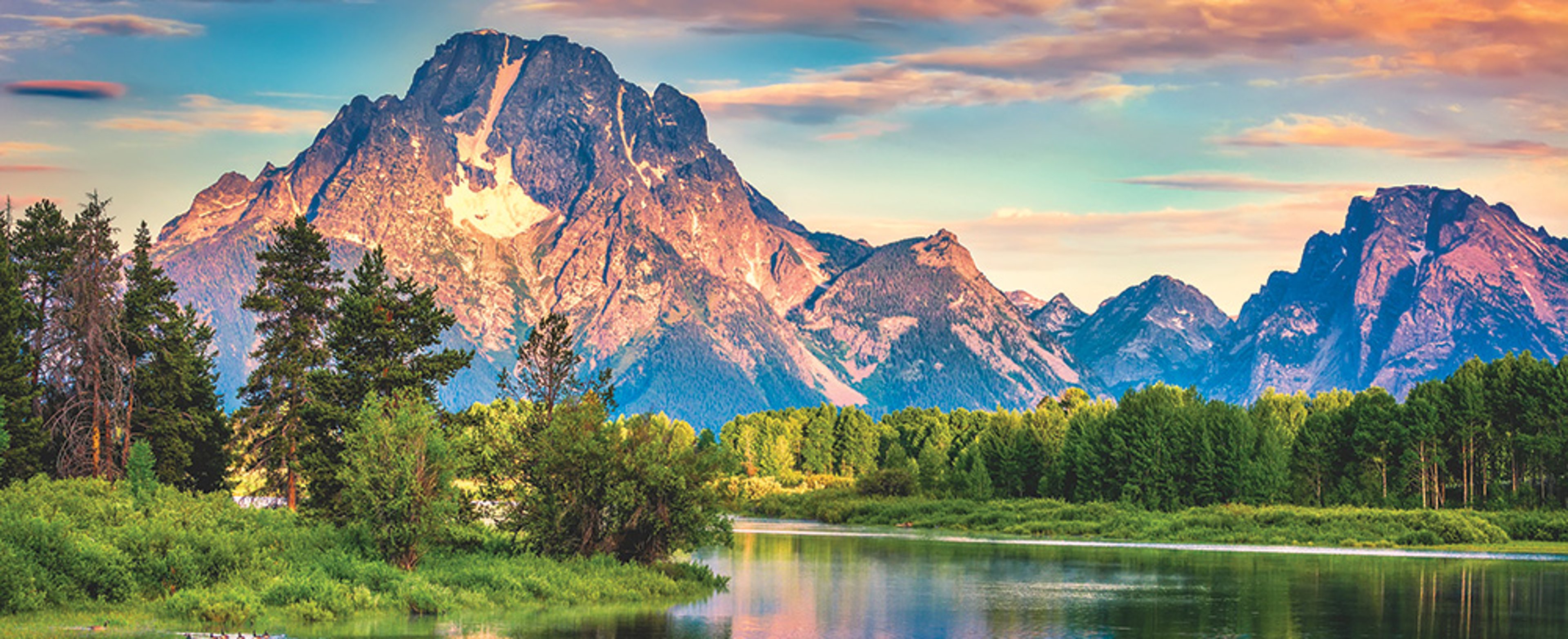 Scenic view of Snake River with lush greenery and a mountain range under a colorful sky in Grand Teton National Park